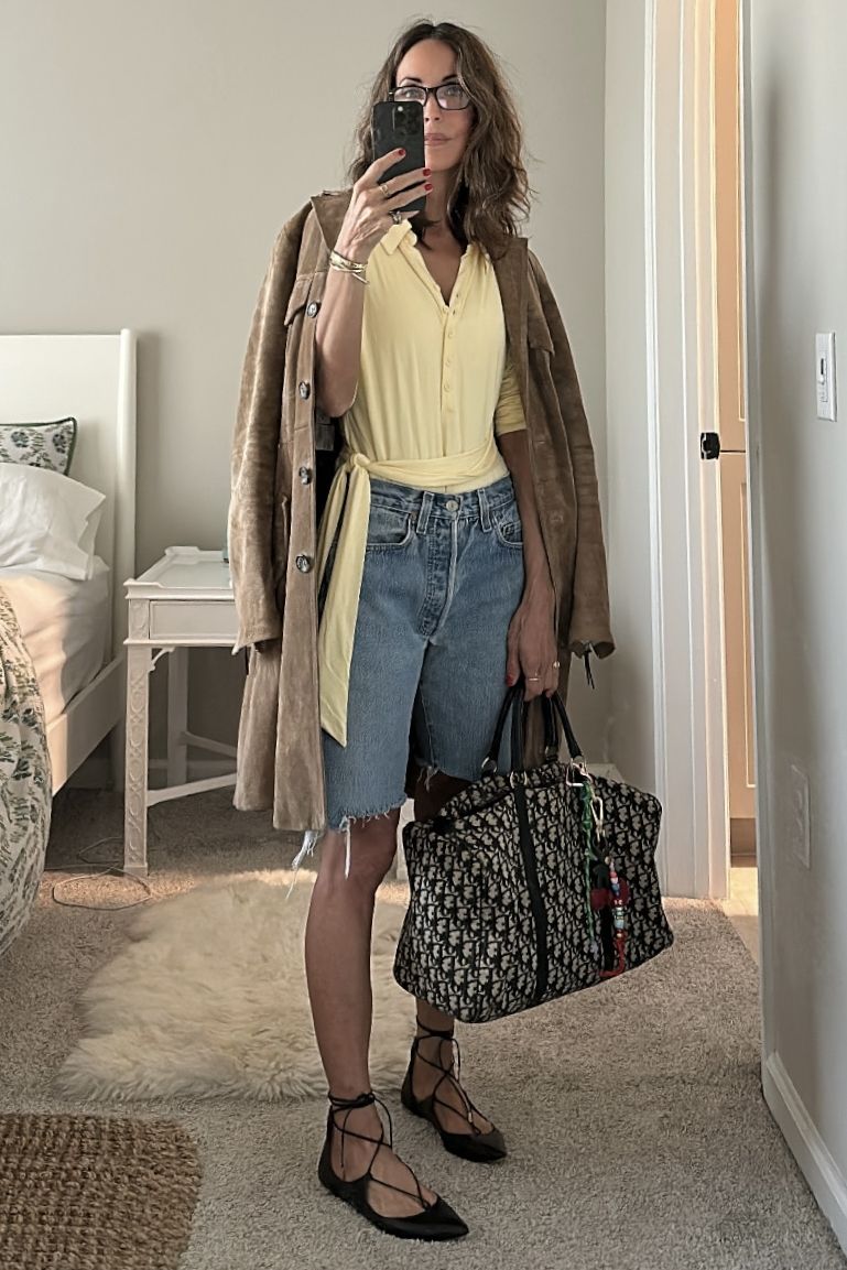 Larissa Mills mirror selfie carrying a Dior duffle bag and wearing blue jean shorts, a yellow shirt, brown coat, and black flats.