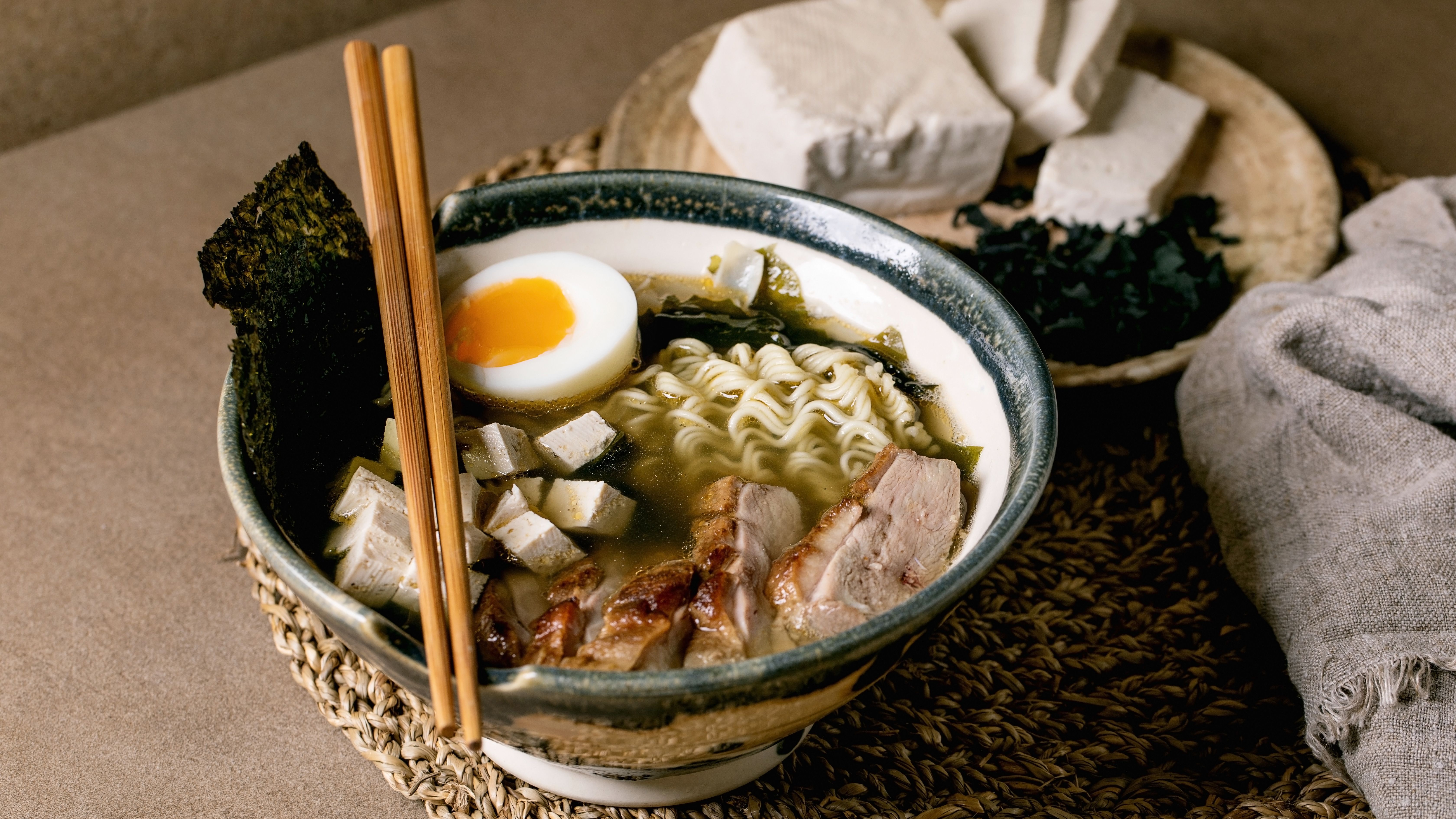 Homemade japanese style soup ramen with noodles, grilled duck breast, tofu, seaweed nori chips and boiled egg in ceramic bowl with chopsticks and spoon on brown table with knitted straw napkin. (Photo by: Natasha Breen/REDA/Universal Images Group via Getty Images)