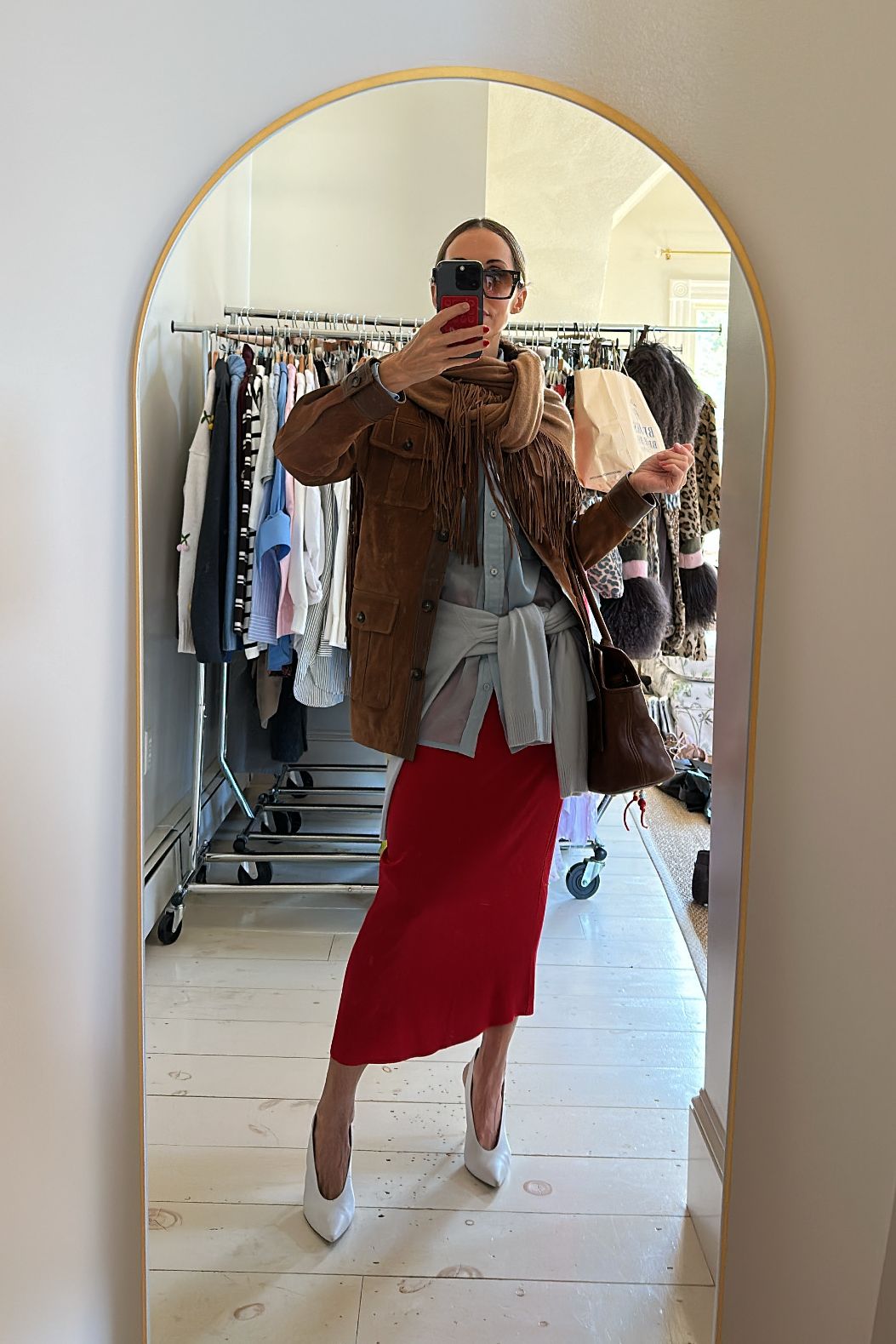 Larissa Mills in a red skirt, blue button-down, blue sweater around the waist, brown suede jacket with fringe, and white heels