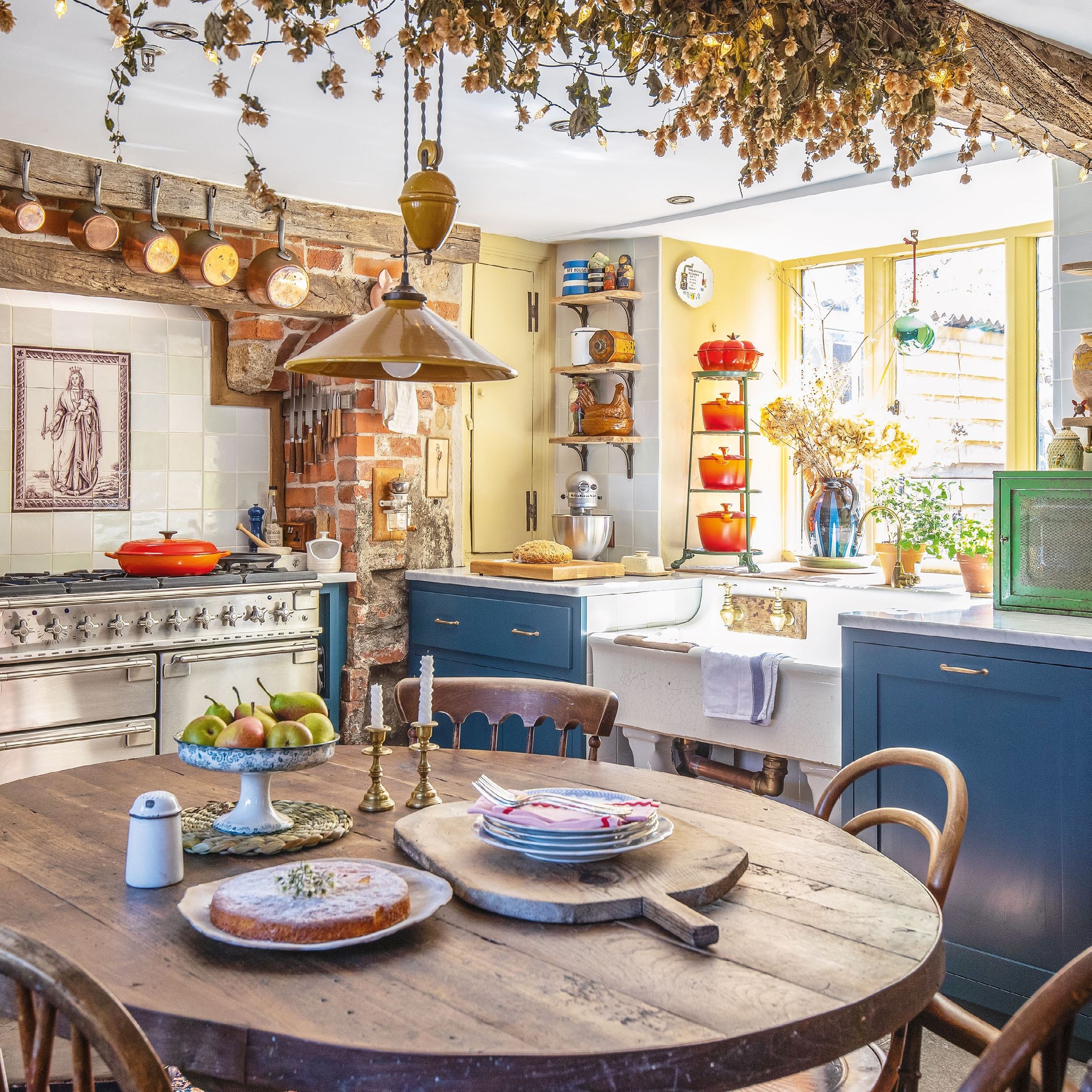 Country cottage kitchen with round wooden table and chairs underneath dried floral arrangement. Blue cabinets and large stainless steel range.