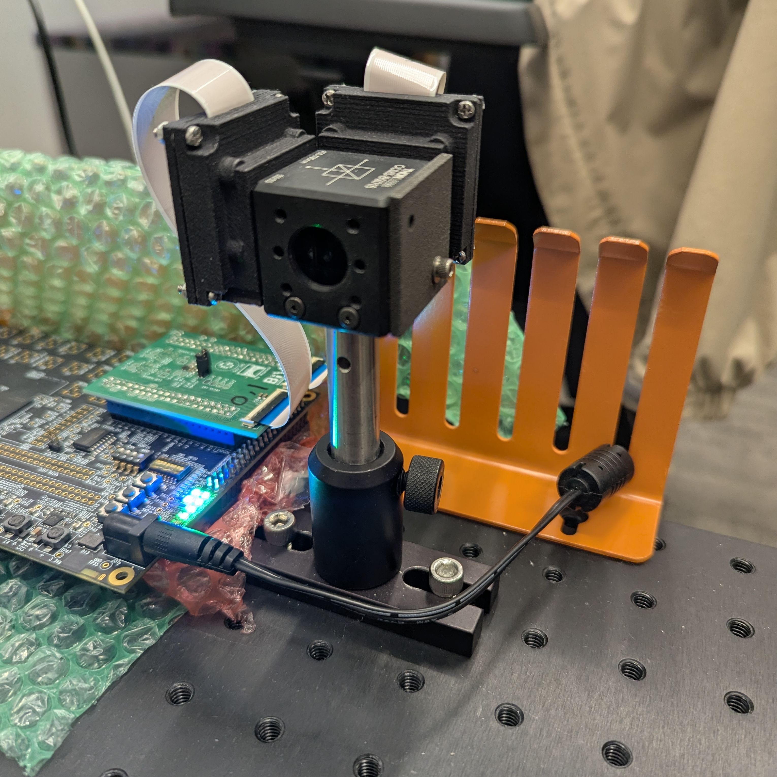 An optomechanical setup featuring a Thorlabs beamsplitter cube is mounted on a metal post above an optical breadboard, flanked by a custom circuit board with green LEDs and an orange component rack.