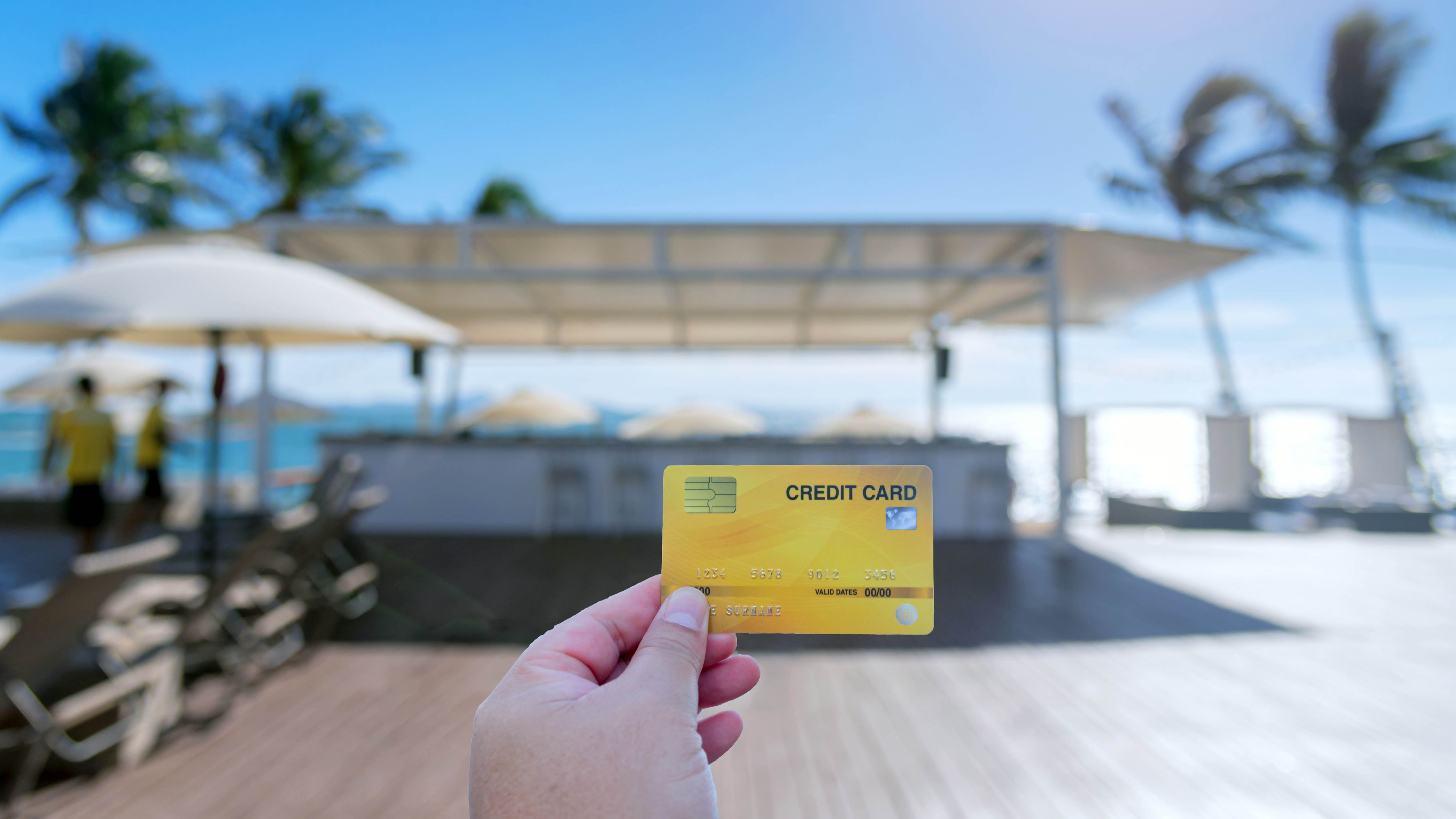 A hand holds a credit card in the foreground while a beach resort bar is in the background
