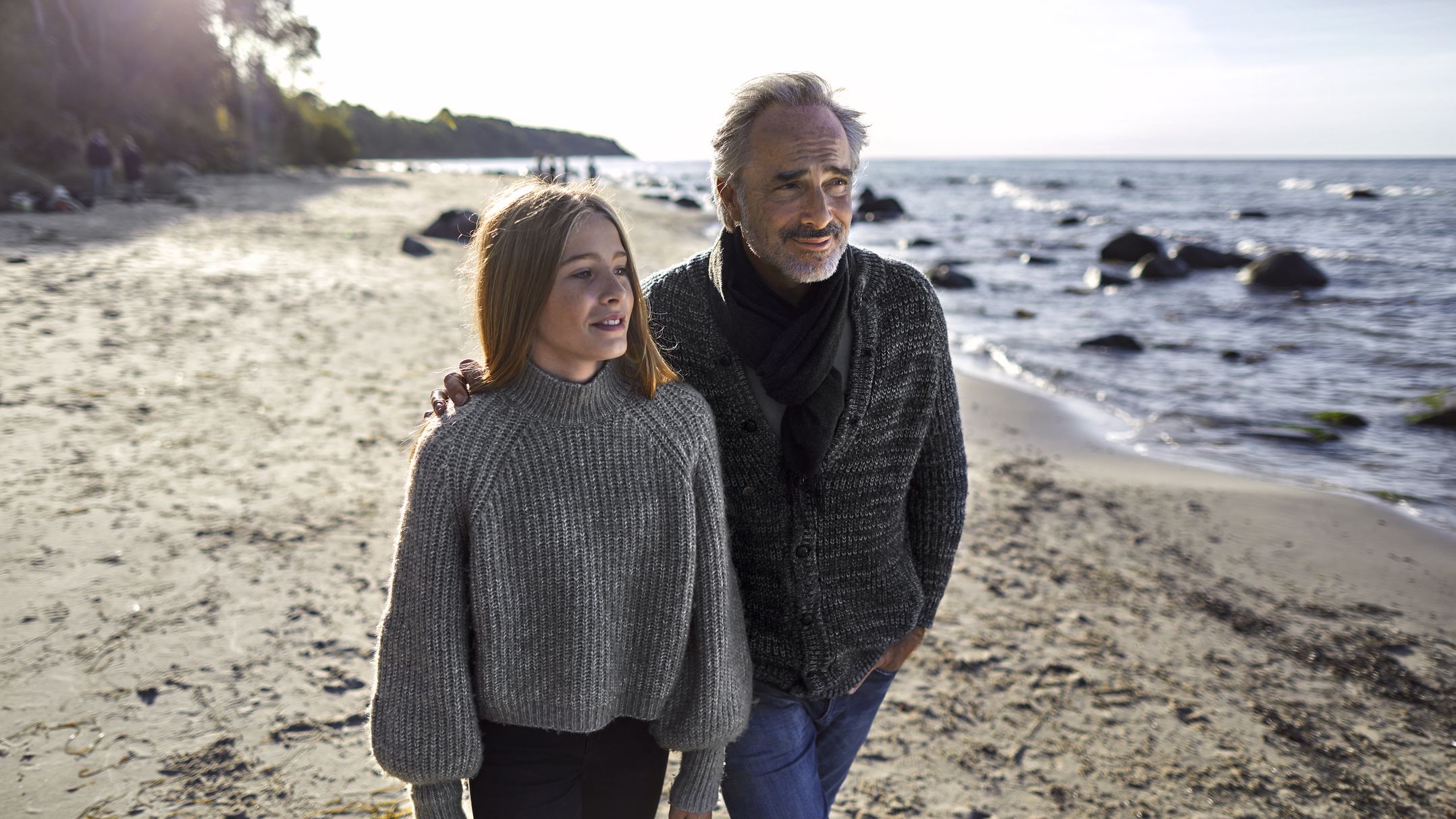 A man and his older daughter walk together on the beach on a blustery day.