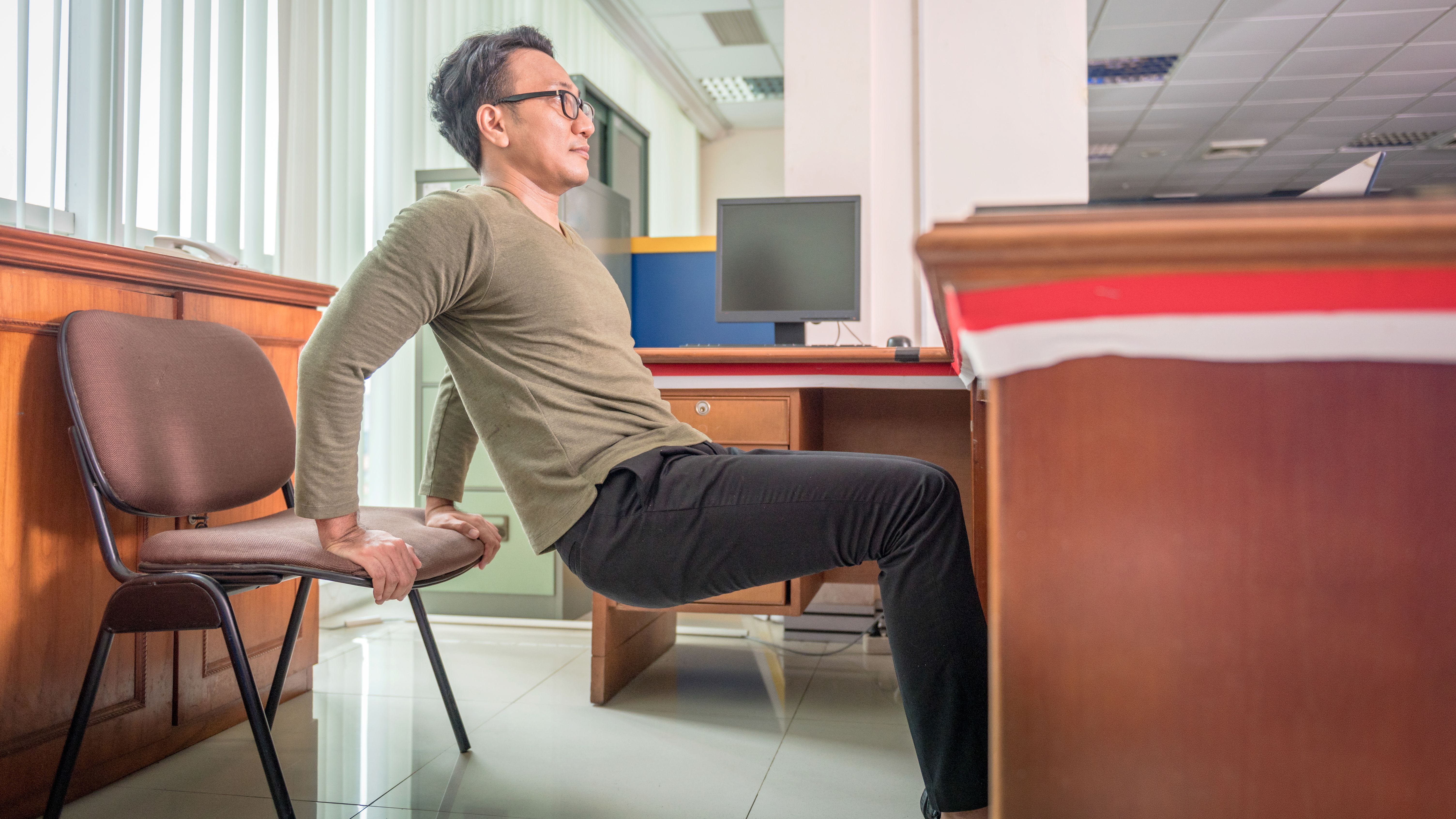 A man practices triceps dips at his desk on a chair. He has pushed the chair back away from the desk; his hands are on the edge of the chair, his feet placed far in front and his knees are bent. In front of him is a wooden desk. 