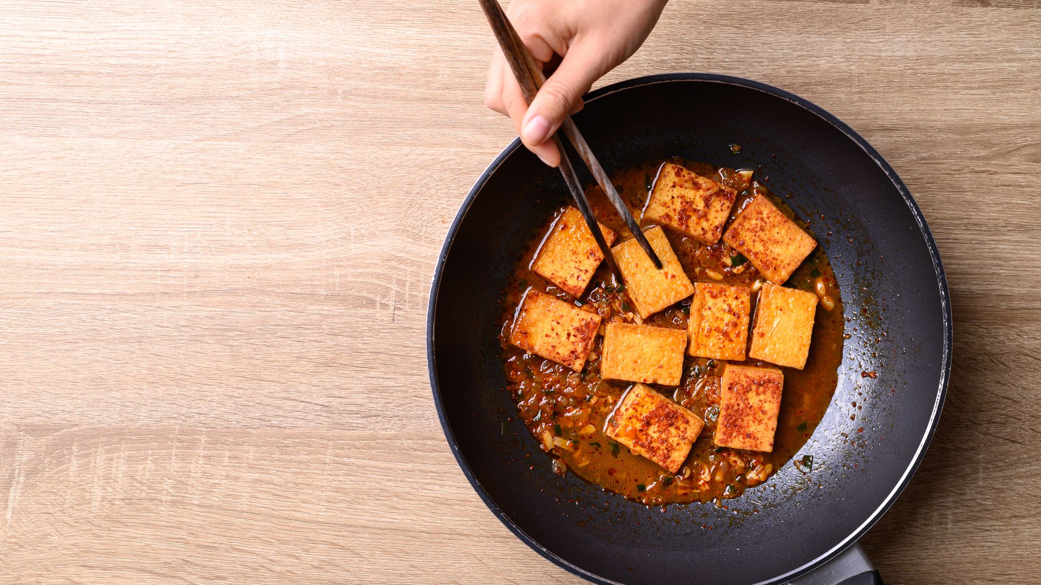 Braised tofu in cooking pan on a tabletop with hand and chopsticks