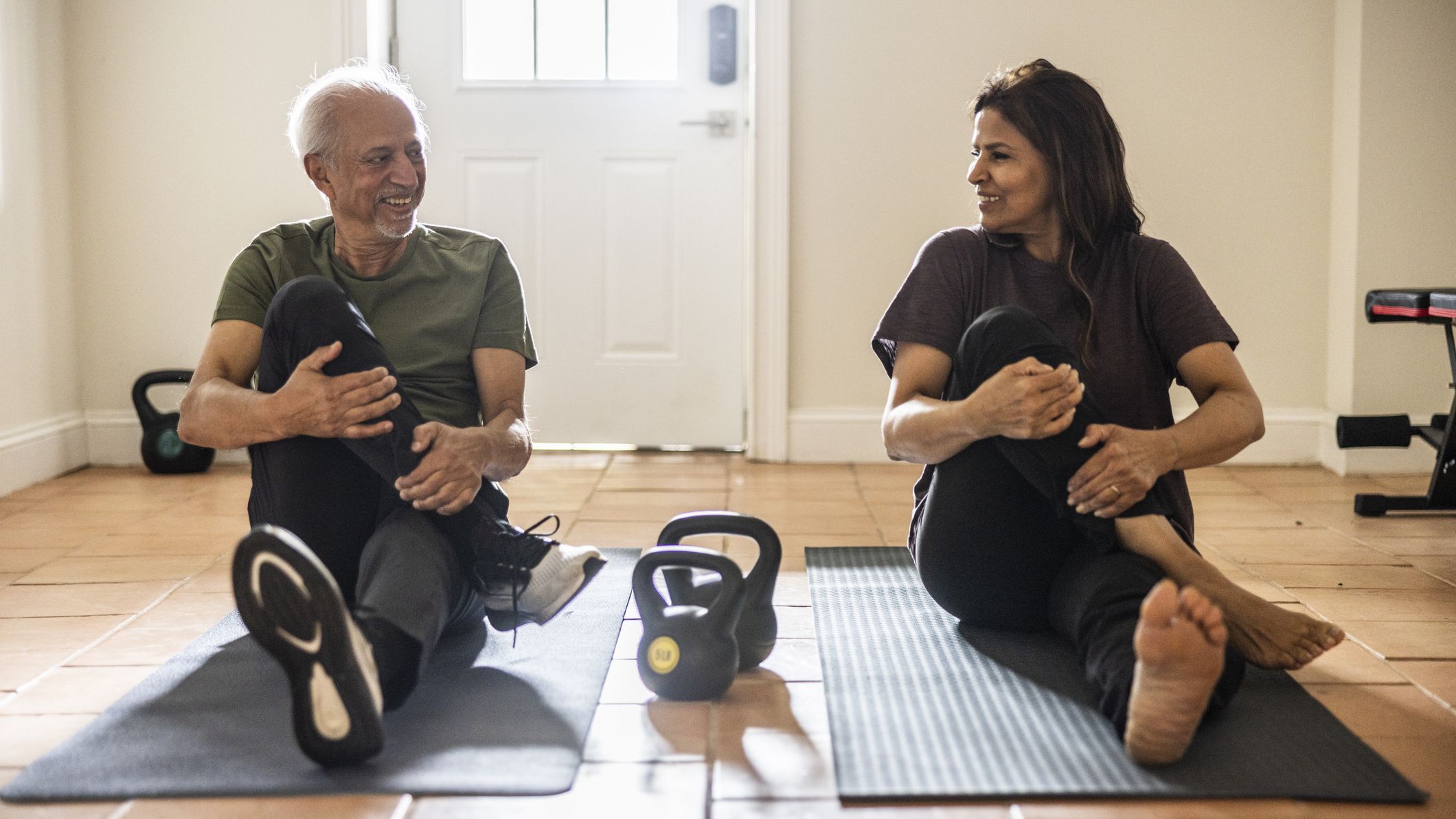Senior man and woman sit on exercise mats performing a glute stretch, with kettlebells on the floor between them