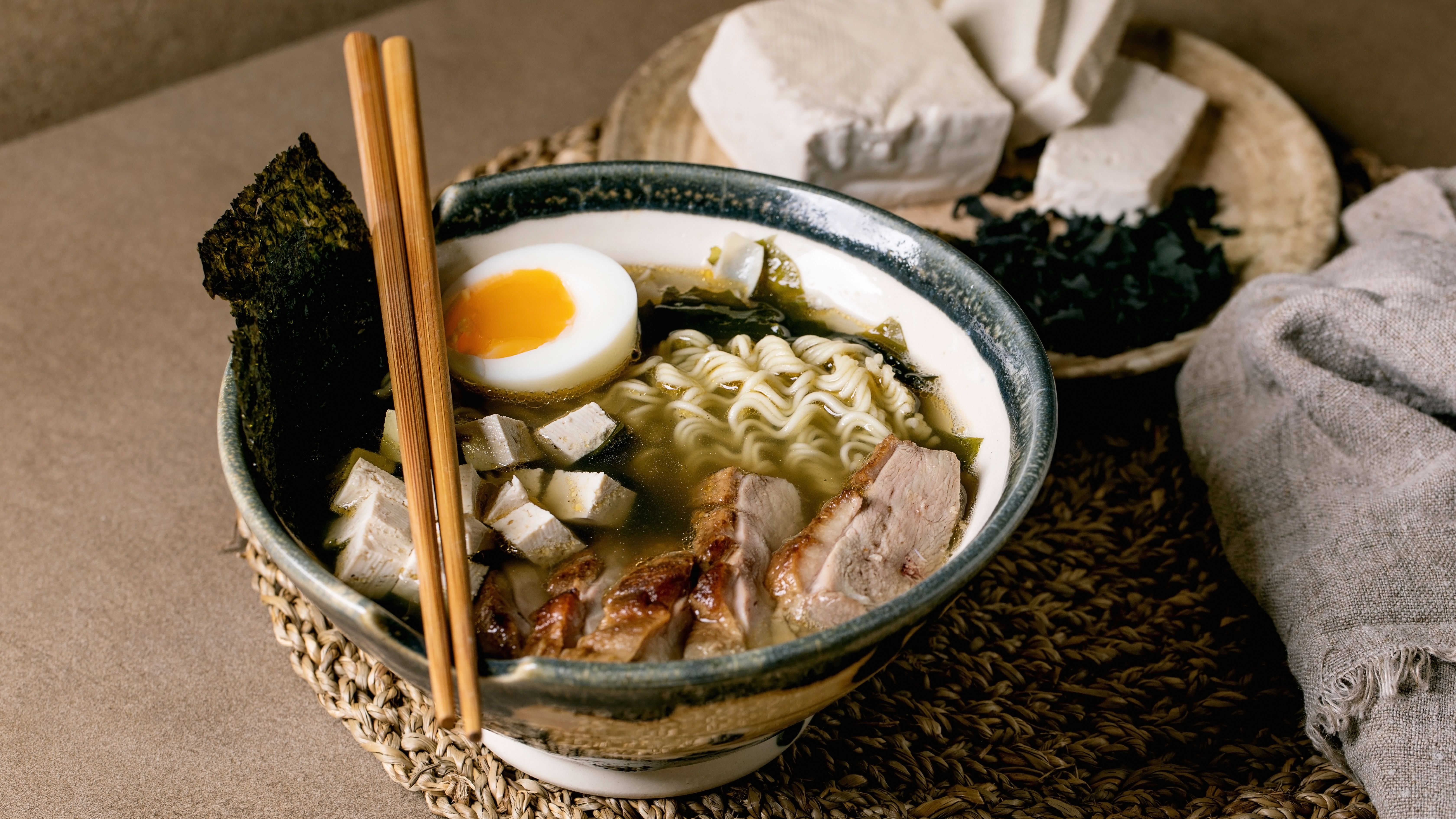 Homemade japanese style soup ramen with noodles, grilled duck breast, tofu, seaweed nori chips and boiled egg in ceramic bowl with chopsticks and spoon on brown table with knitted straw napkin. (Photo by: Natasha Breen/REDA/Universal Images Group via Getty Images)