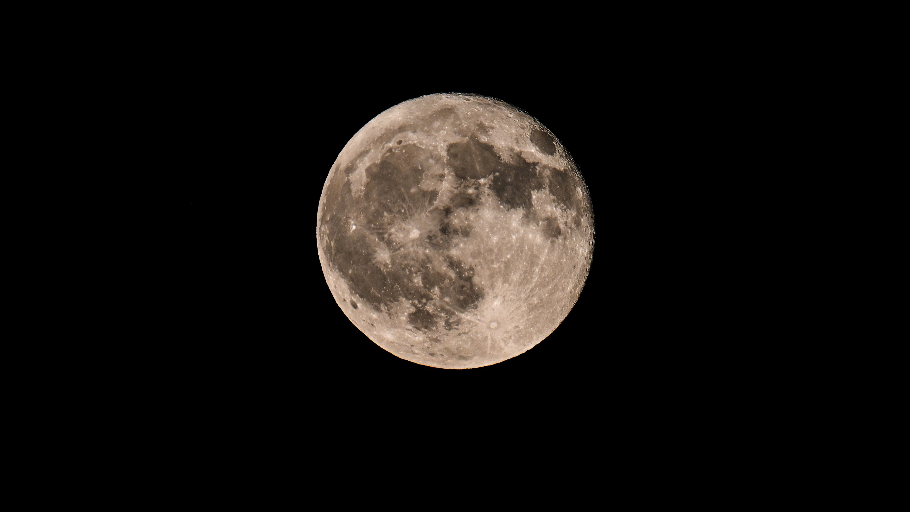 Uma lua cheia é fotografada em um céu noturno negro com mares lunares escuros e crateras marcando sua superfície.