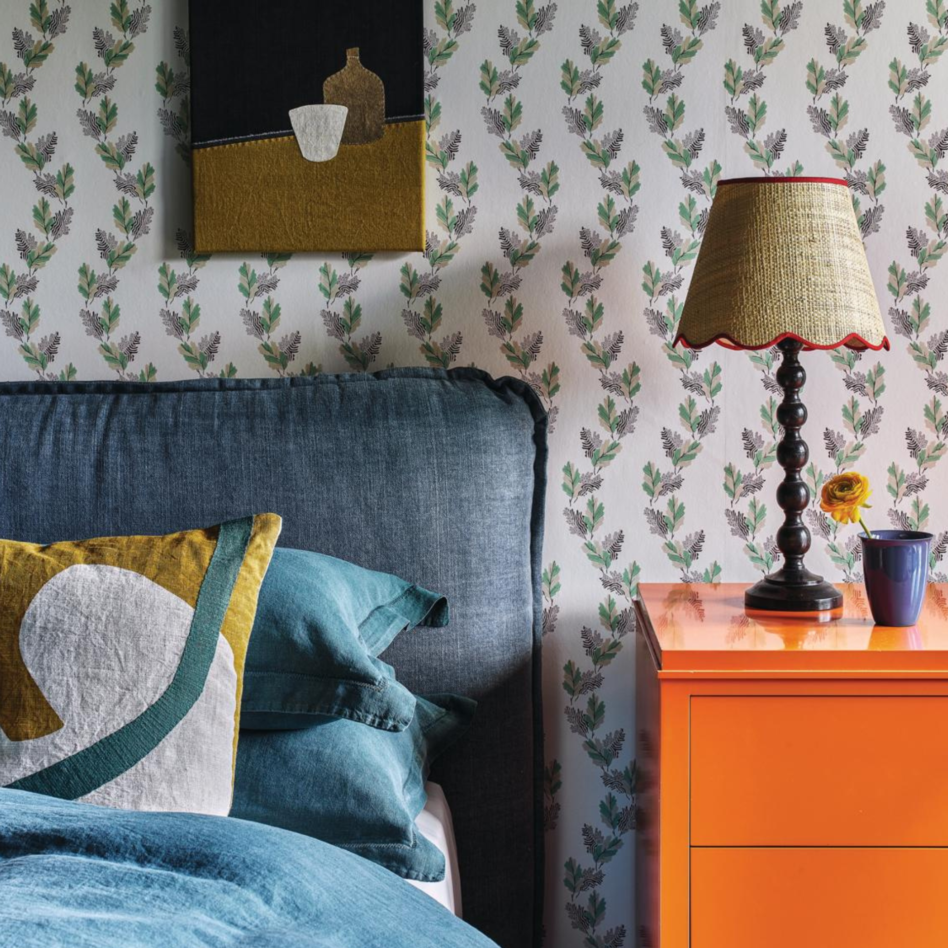 a bedroom with floral vine wallpaper, a bed with a blue headboard, and a bright orange side table