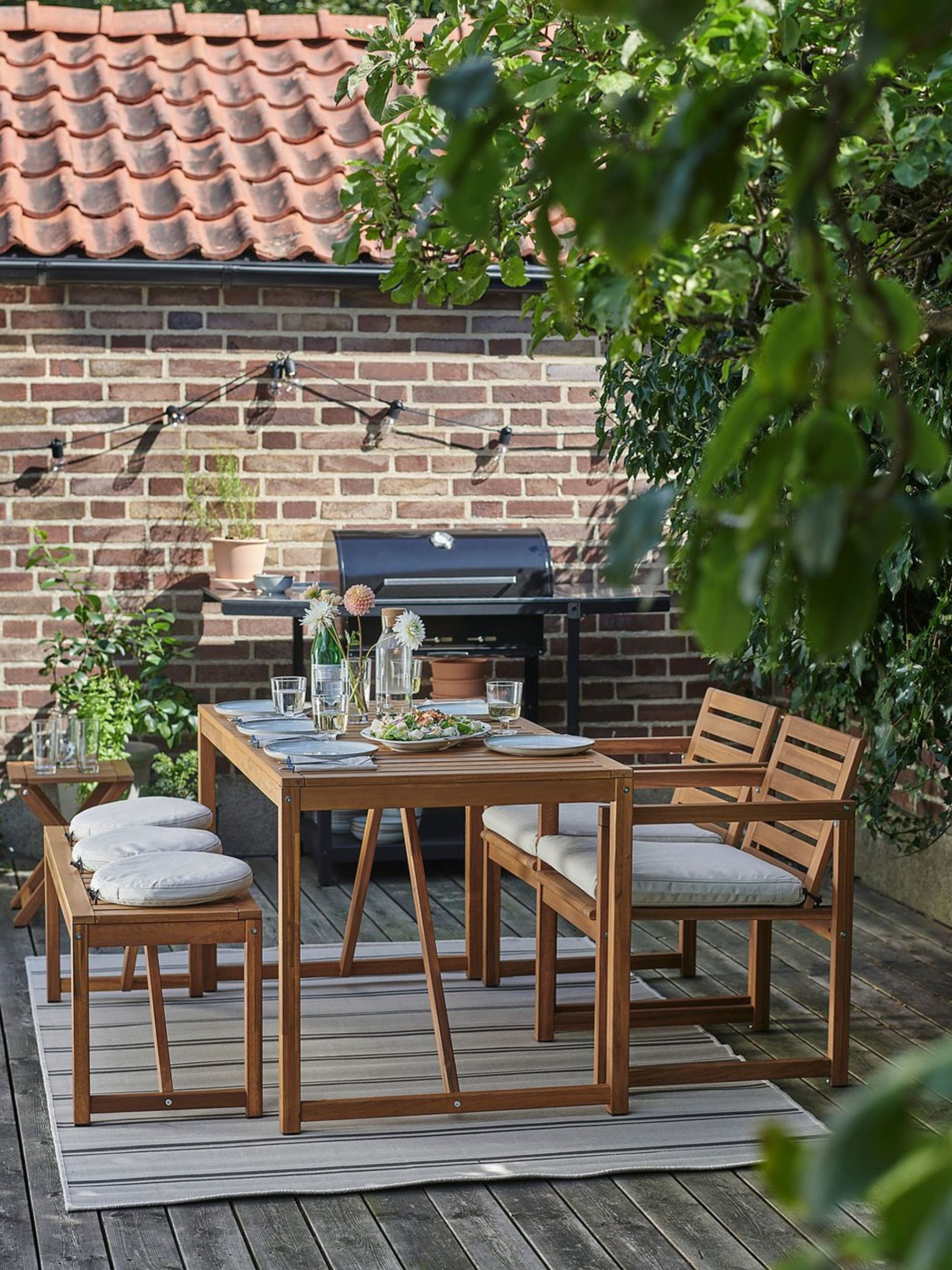 An outdoor wooden dining table with two chairs and a bench with cushions sits on top of a rug on a deck set for a meal with a BBQ in the background and a set of black string lights on the wall