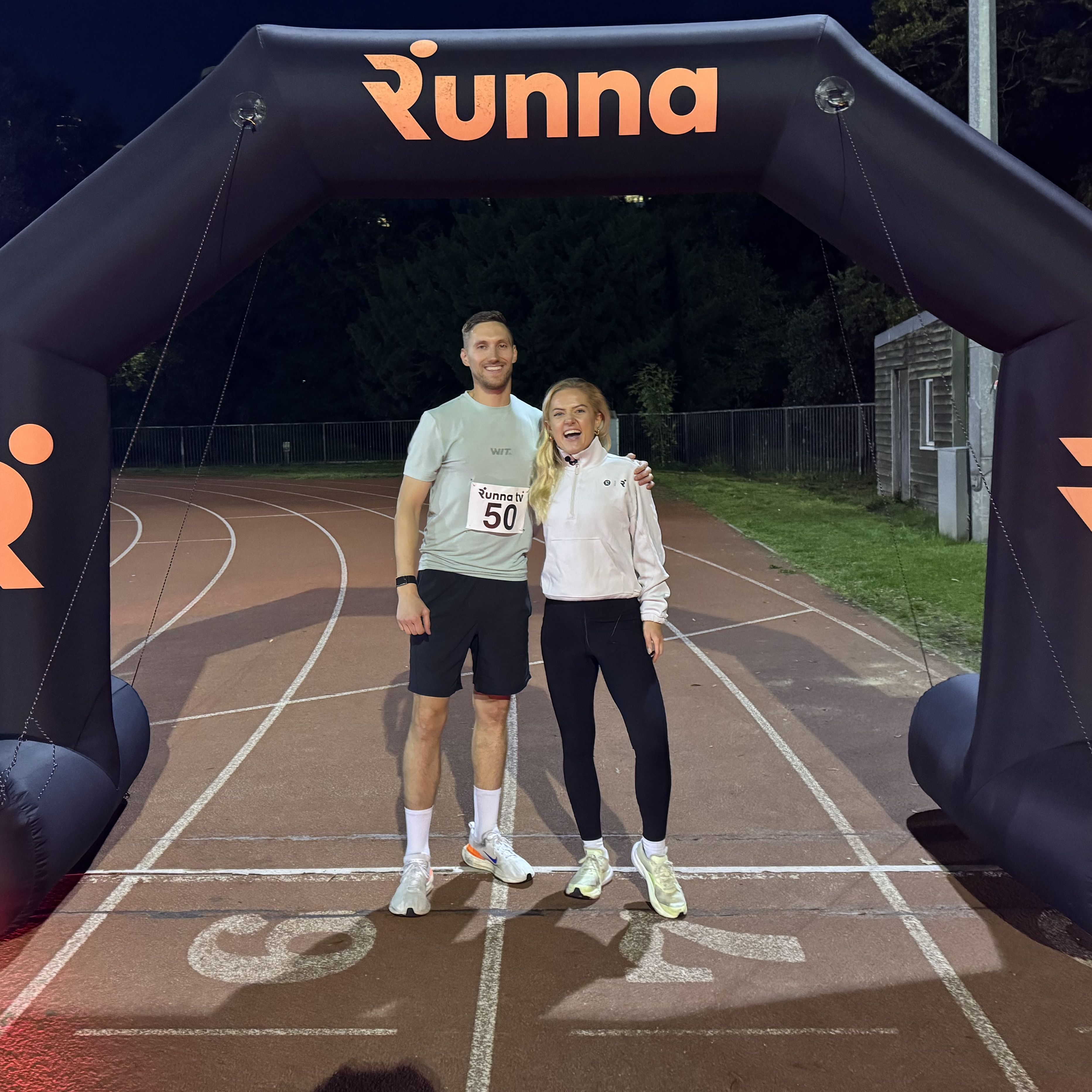 Man and woman on running track standing under black inflatable arch with Runna written on it in red letters