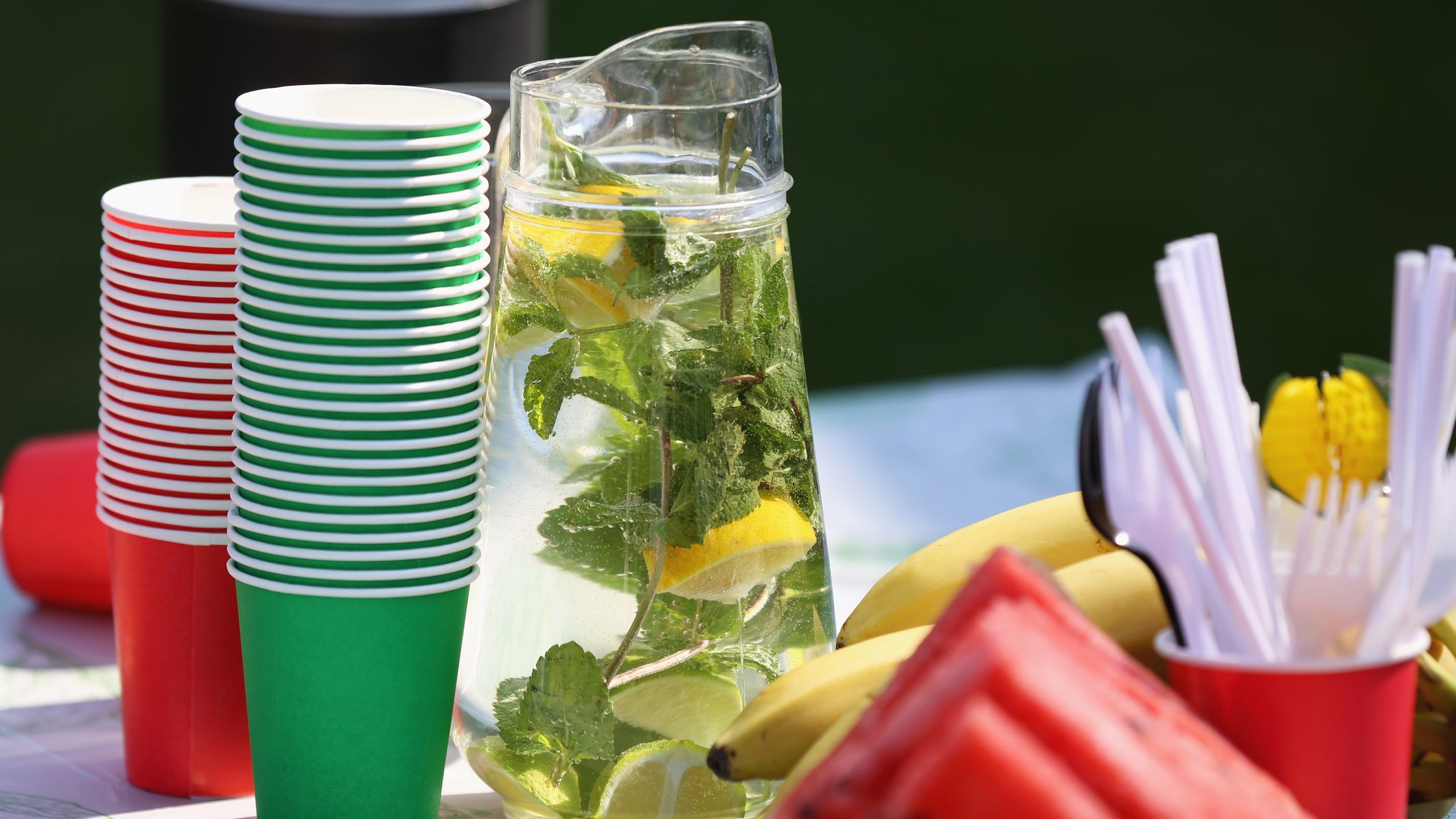 A pitcher of fresh lemonade on a table with plastic cups and watermelon.