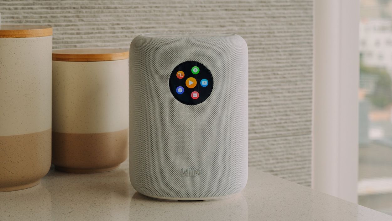 A white WiiM Sound smart speaker on a white surface in front of some jars.