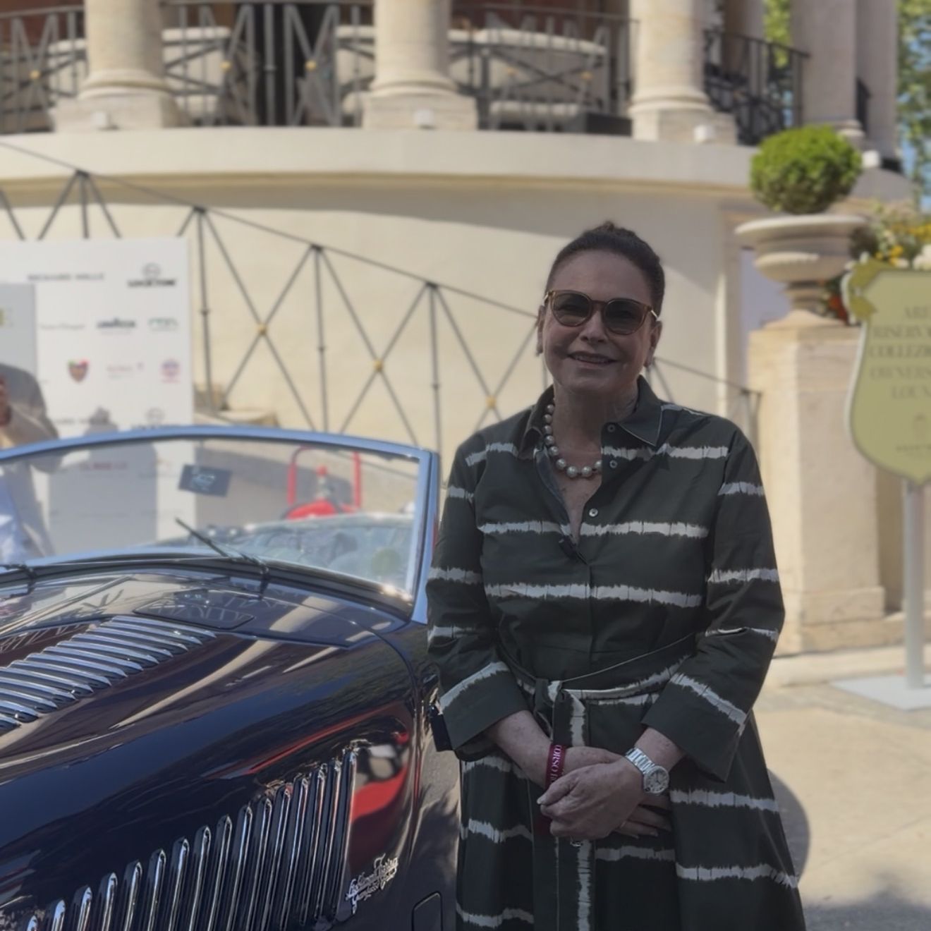 A woman in a striped dress stands beside a dark blue vintage convertible outside Casina Valadier during Anantara Concorso Roma, with the villa&amp;rsquo;s colonnaded terrace behind her.