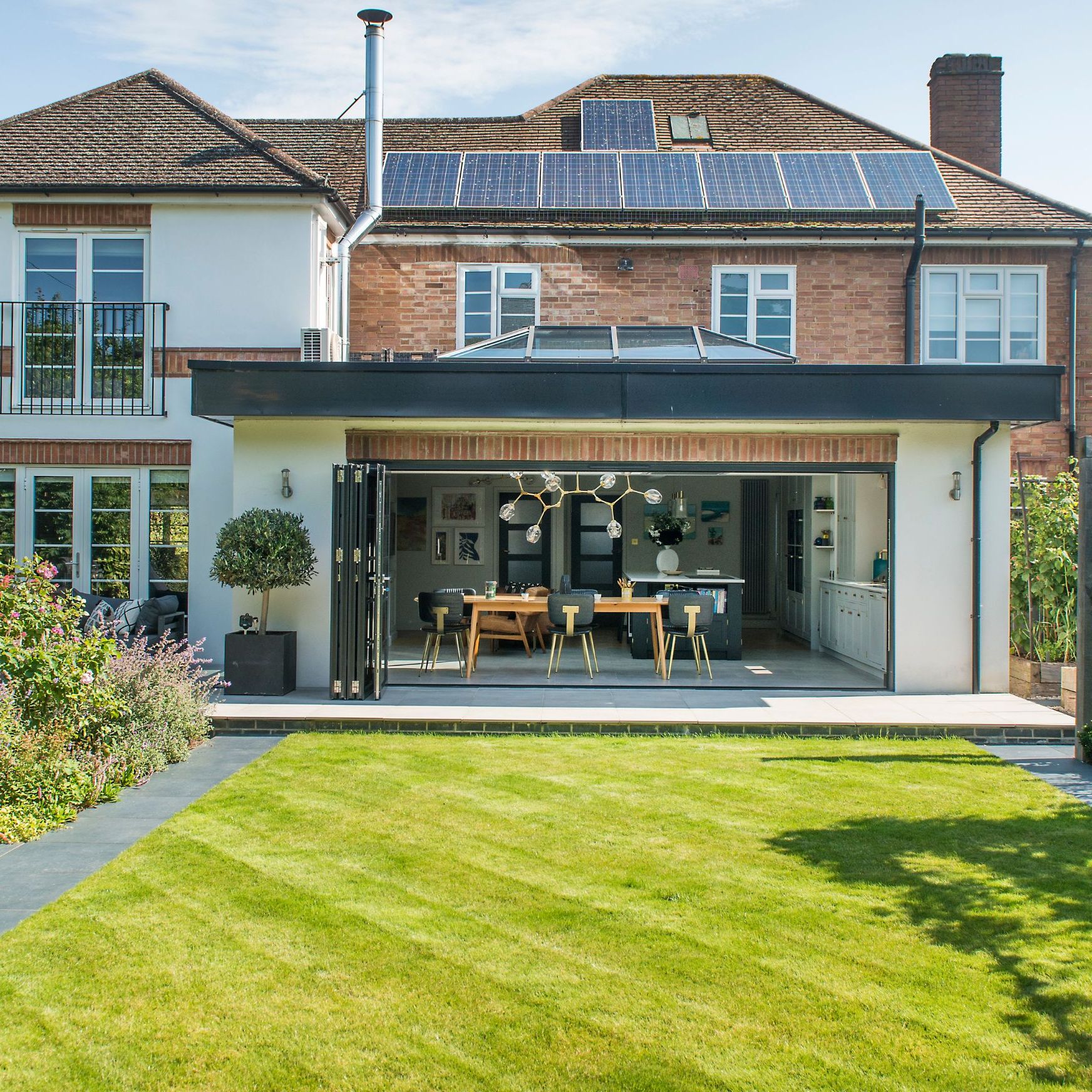 Rear elevation of large home with solar panels on the roof, a roof lantern on a flat roof extension, green lawn and plush garden