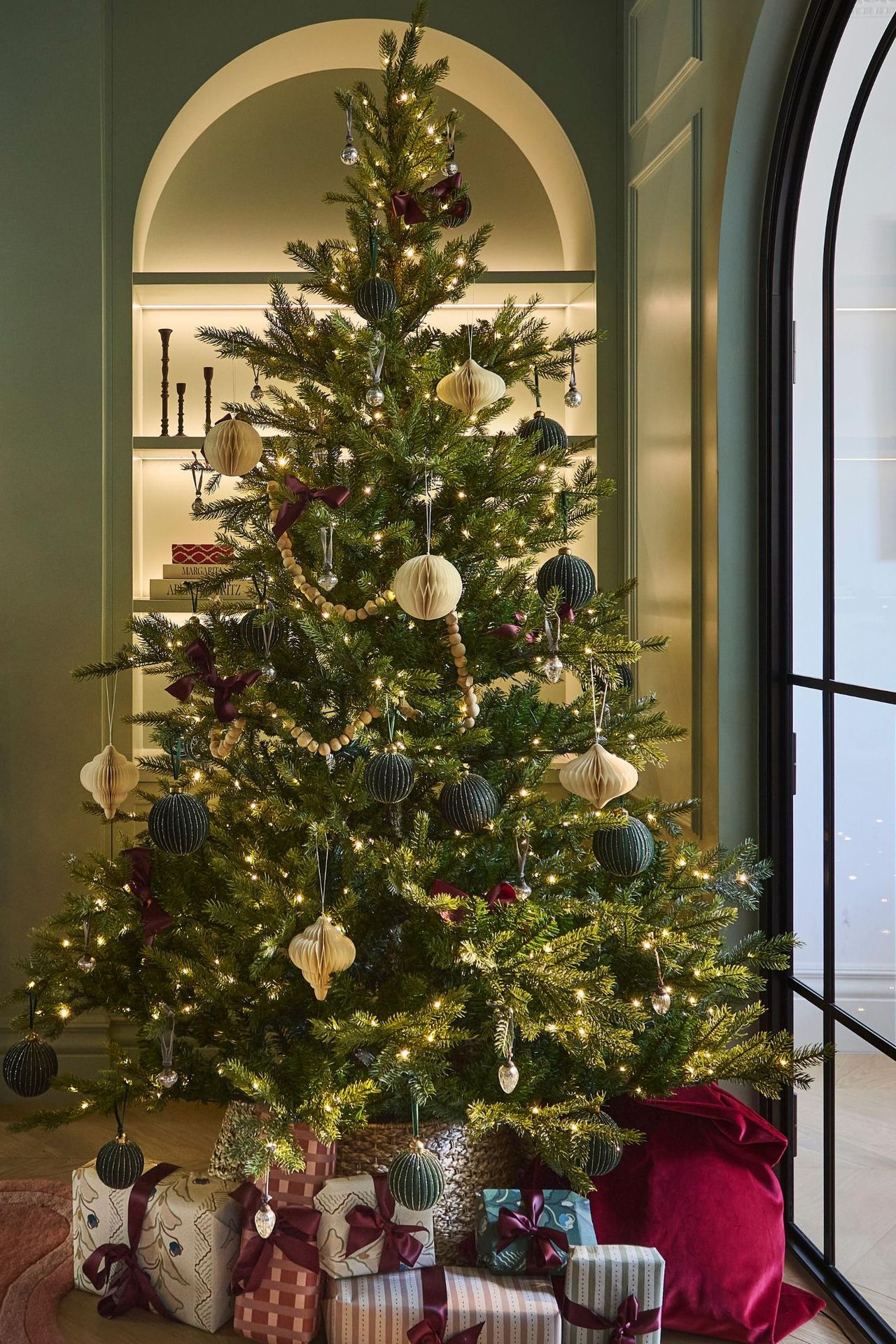 a muted green victorian living room with a marble fireplace, TV, arched bookcase all decorated for Christmas with a large lit tree and wrapped gifts