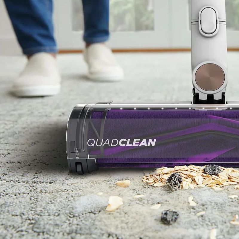 Close-up of a white and purple Shark vacuum floorhead vacuuming unidentified dust and dirt from gray carpet, with blurred-out person&#039;s legs and feet behind.