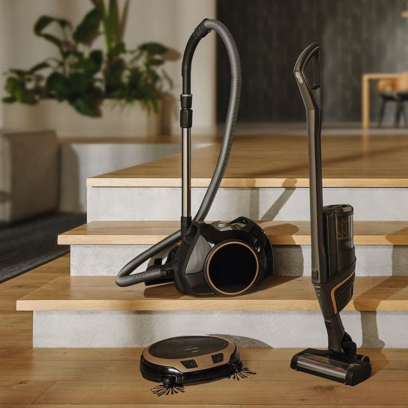 Four Miele vacuum cleaners &amp;ndash; a cordless vacuum, robot vacuum and two canister vacuums &amp;ndash; on set of wooden stairs in luxury home, with plants and furniture pictured in background.