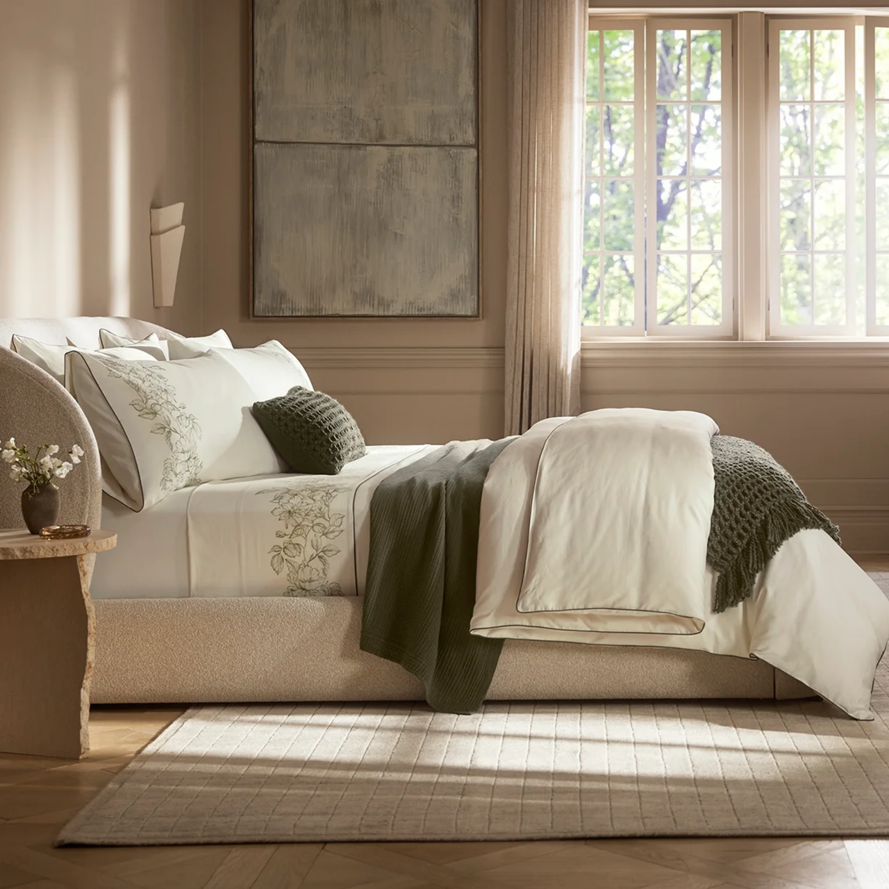 Green and cream colored bed sheets styled in a beige bedroom that features large windows and contemporary art