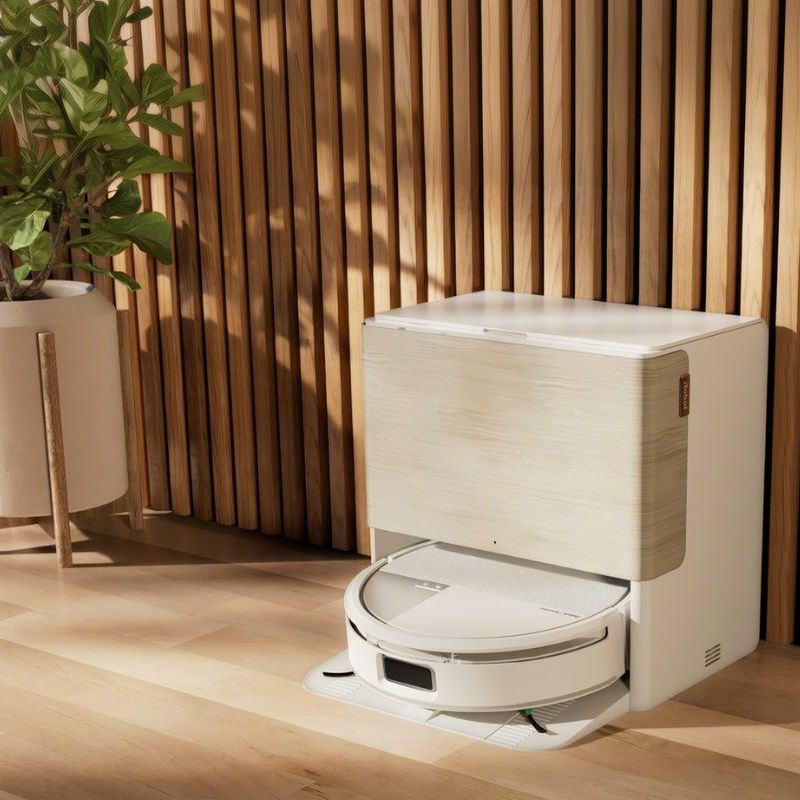 Lifestyle shot of iRobot Roomba Max 705 Combo in stylish living room on a wooden floor, in front of a wooden-paneled wall. A potted indoor tree and drawers are pictured to the left of the white robot vacuum docked in its cleaning station.
