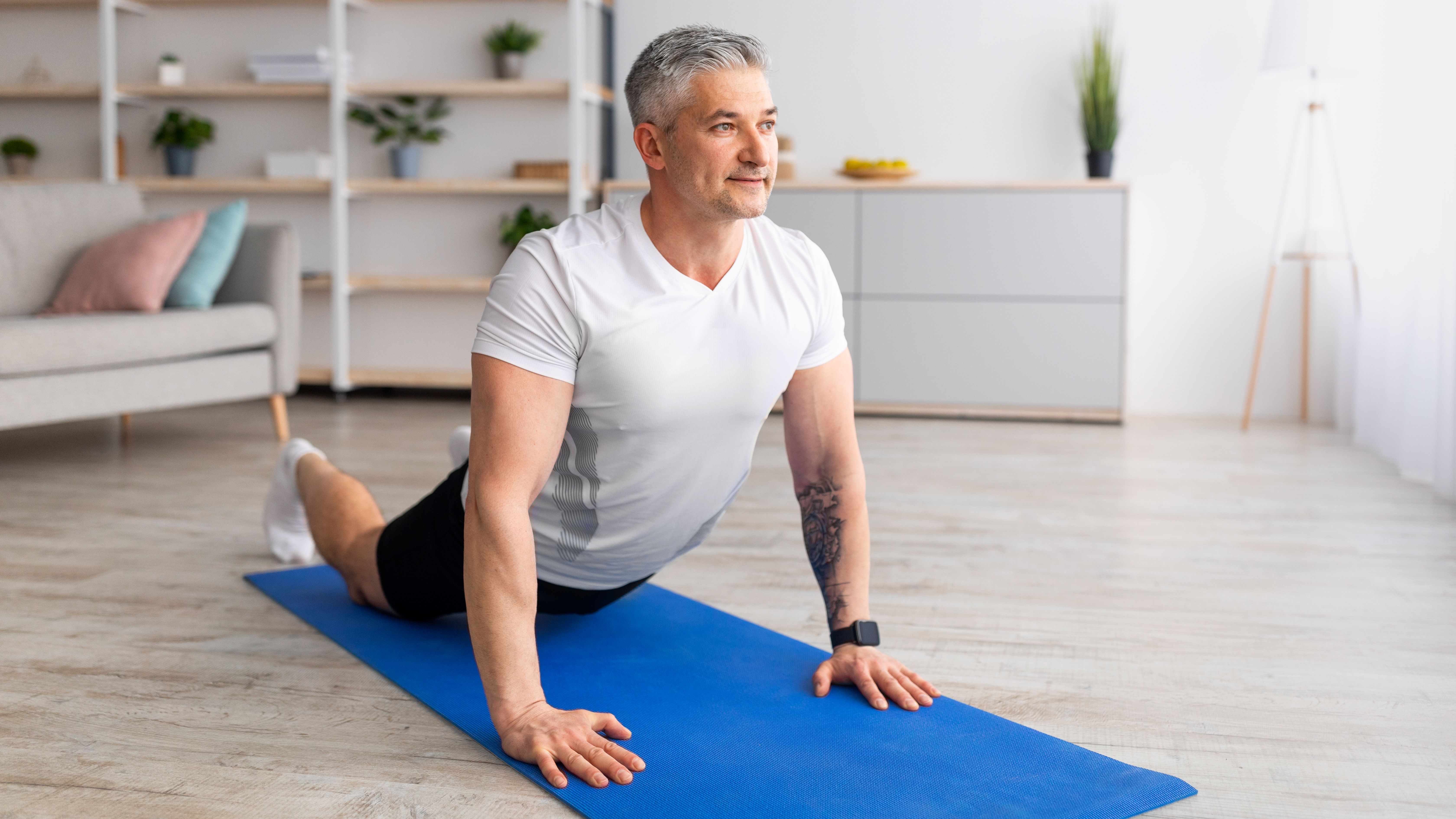 A man practices cobra pose at home on a yoga mat. He is on his front, with his legs long on the mat and his torso held upright by straight arms. Behind him we see a couch and shelves with plants.