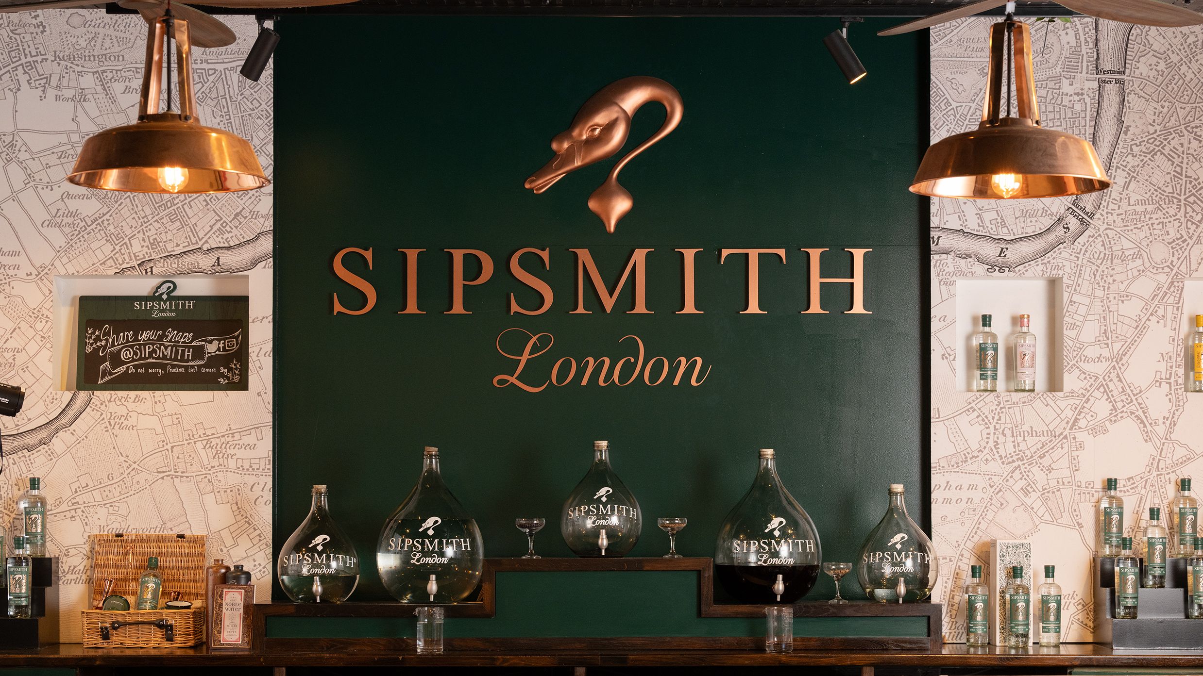 an image of a countertop inside the Sipsmith distillery, with various shaped decanter-style dispensers across the back, and a large Sipsmith sign on the wall