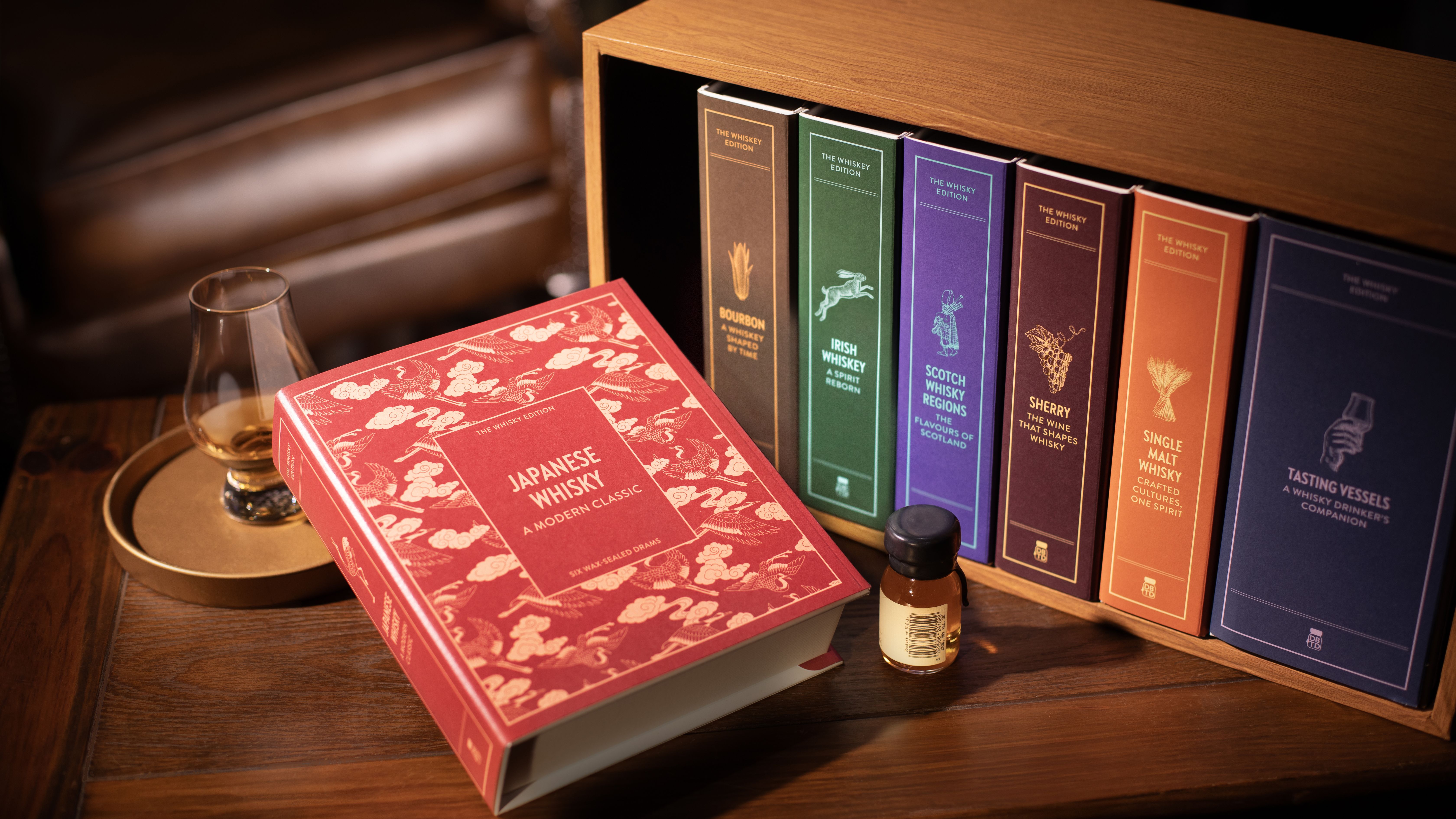 an image of the bookshelf from above with the book on Japanese Whiskey pulled out and laying on a table. Next to it are a tasting glass with a small dram of whisky in it and a sealed miniature