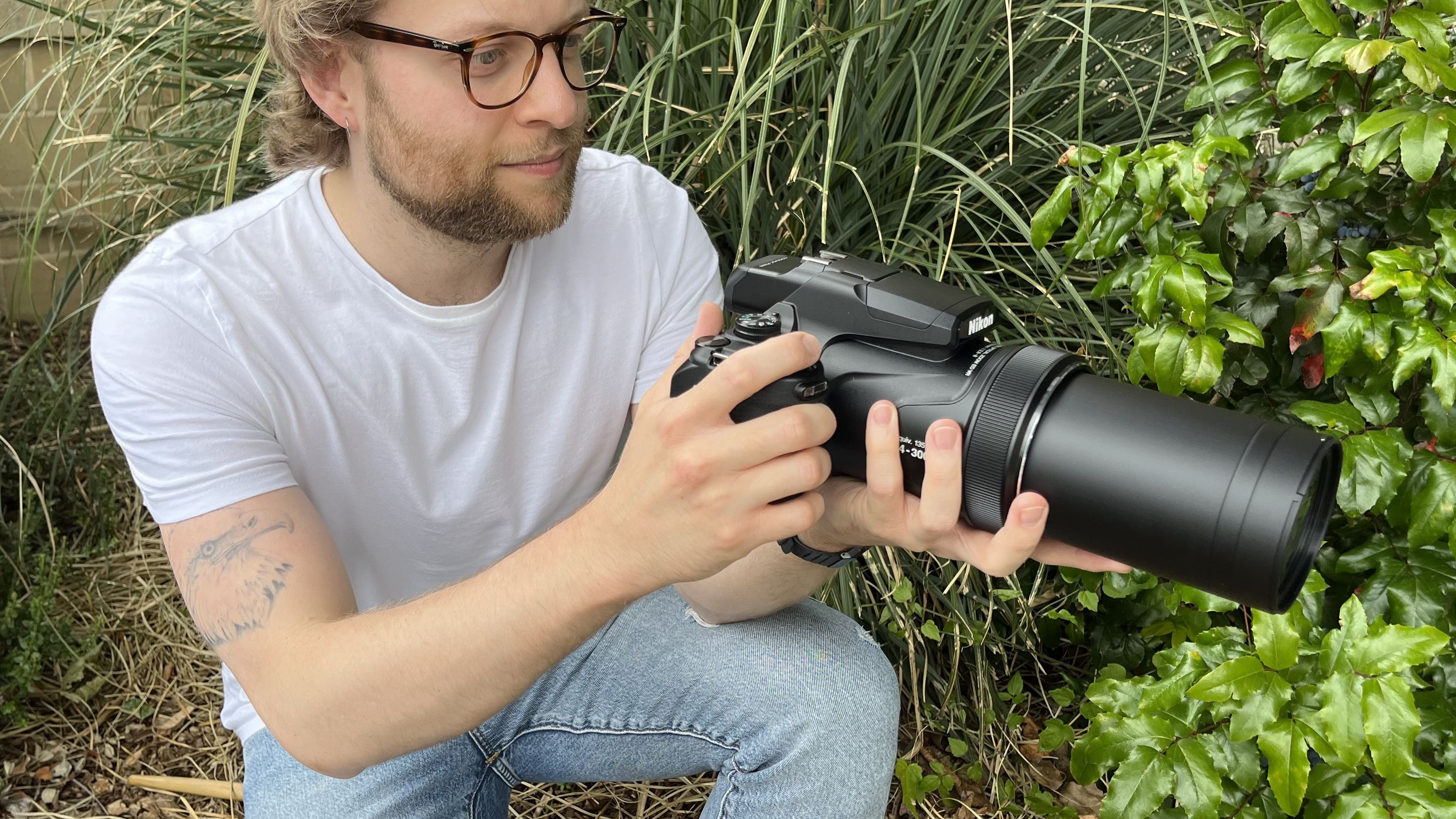 Nikon Coolpix P1100 lifestyle image of crouching Mike Harris holding P1100 in front of vegetation