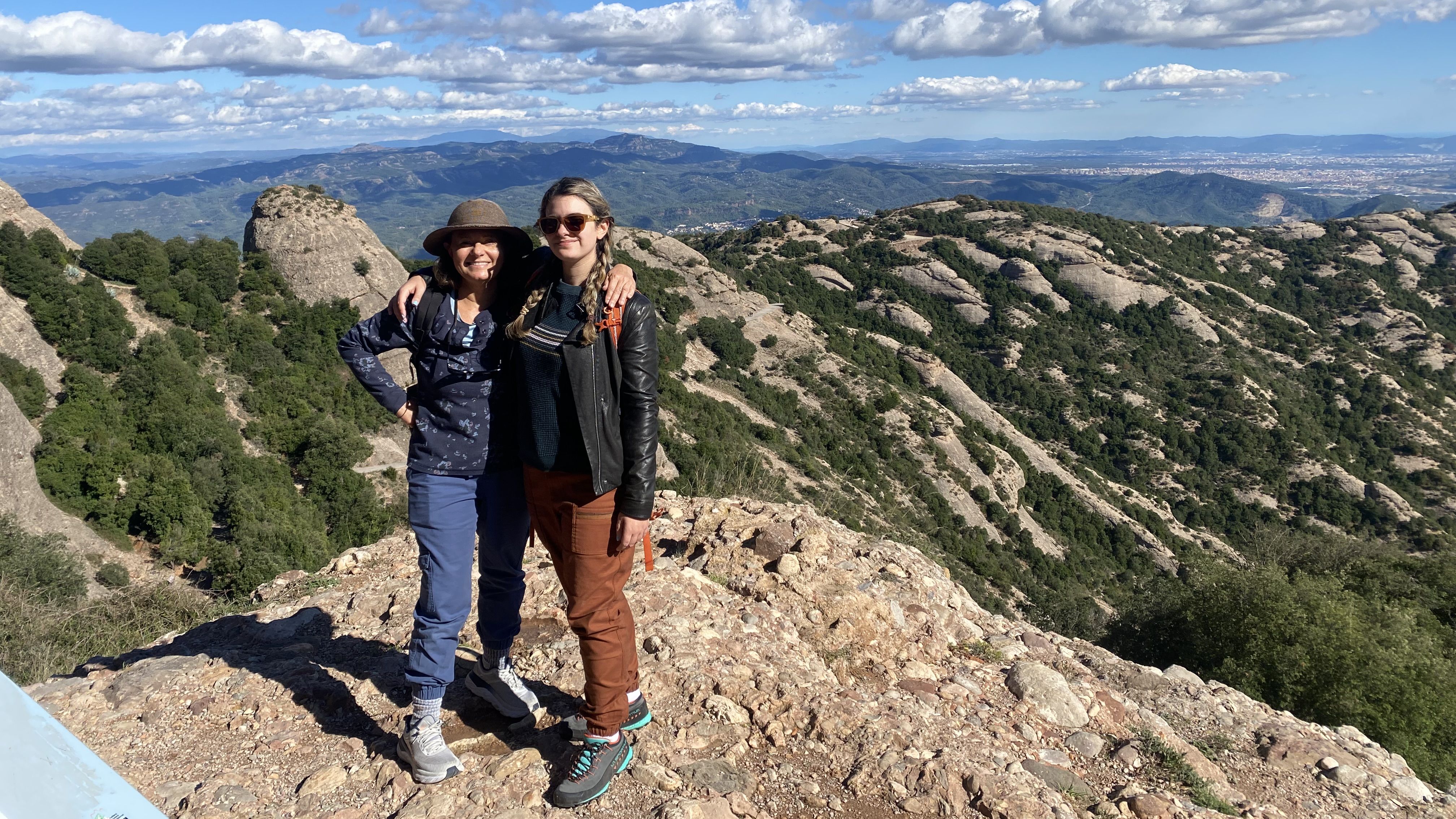A young woman and her mother in hiking clothes stand on a rocky outcrop in Montserrat, Spain.