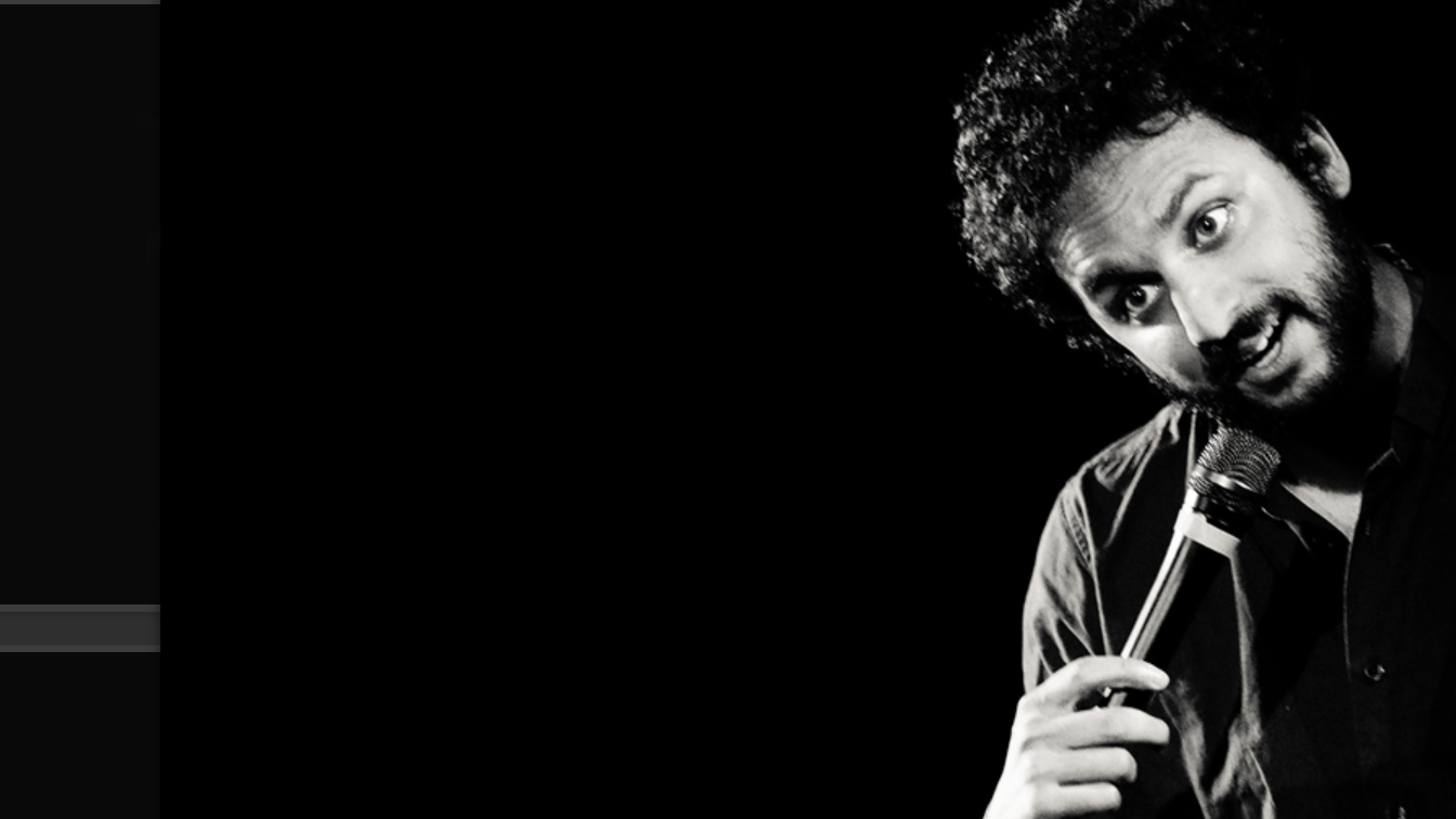 a black and white image of Nish Kumar performing on stage holding a microphone