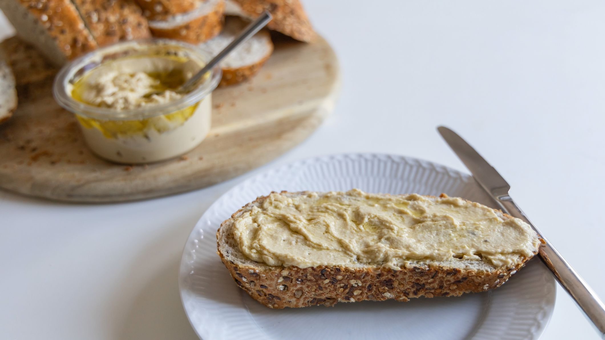 A slice of bread spread with hummus on a plate with a knife resting on the side of the plate, the loaf and tub of hummus are in the background