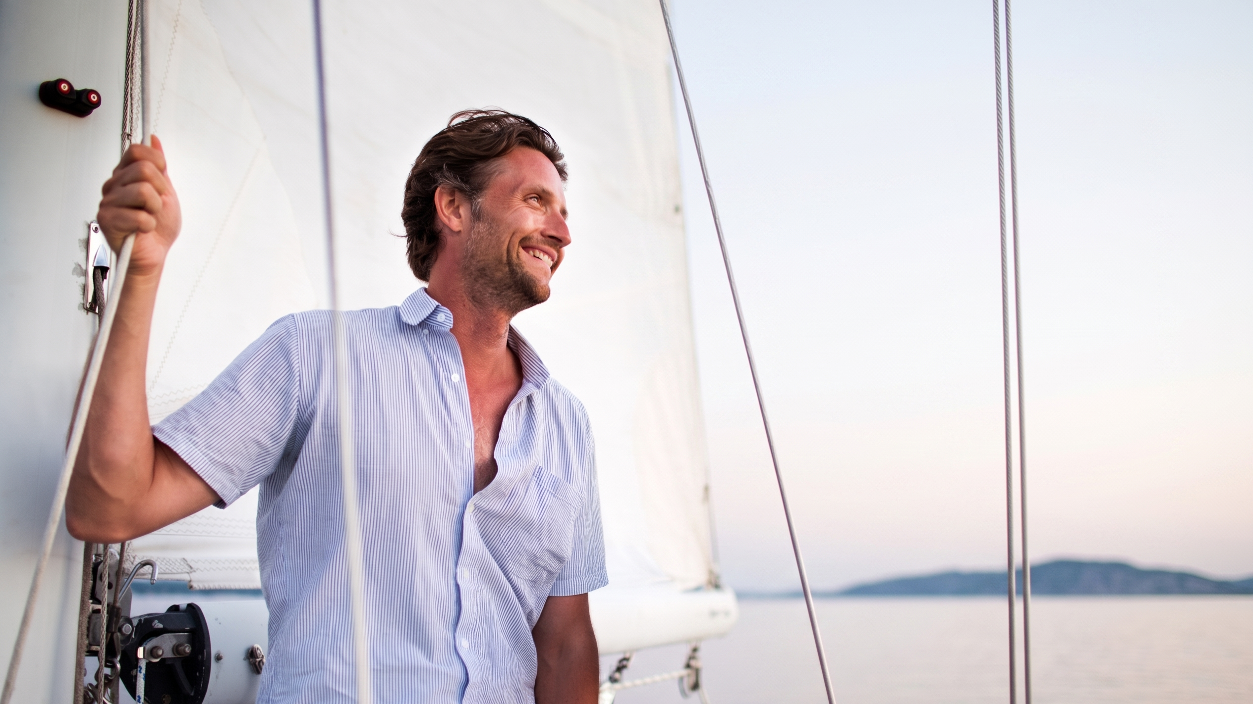 A man in his 50s relaxes on a a sailboat at sunset. he is grinning.