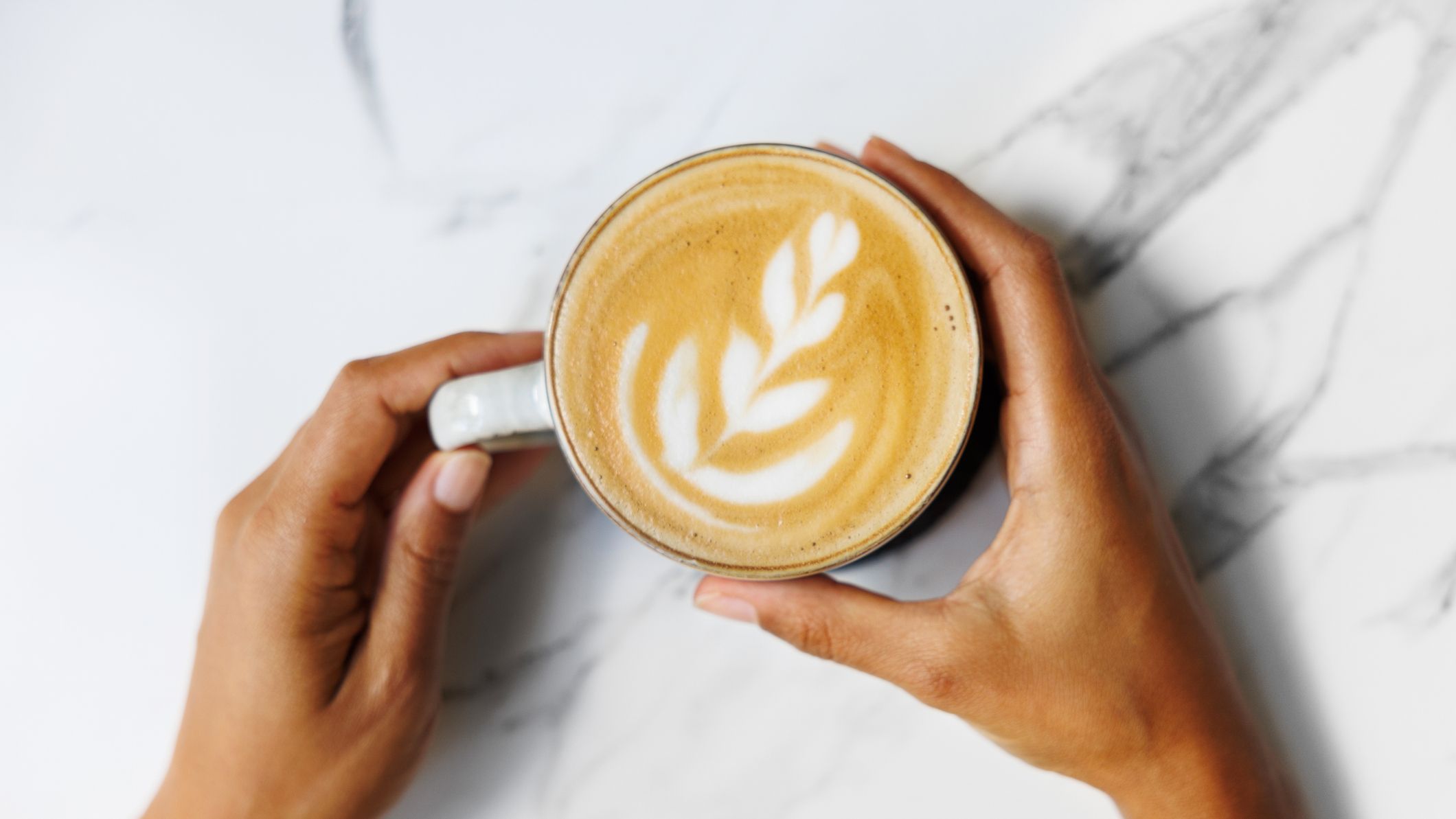Hands gently holding a freshly brewed cup of coffee with intricate latte art on a marble surface.