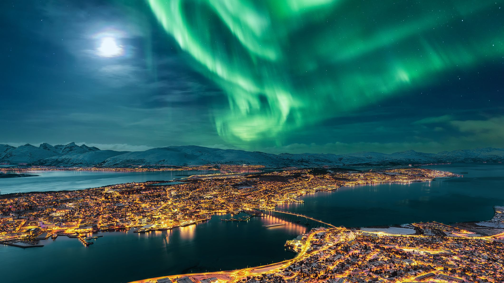 tromso city light shine below a vibrant green aurora and full moon in the upper left.