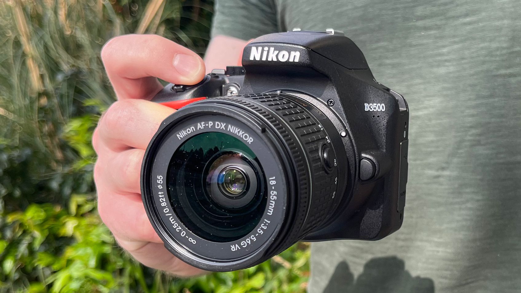 Nikon D3500 being held by person against background of green foliage