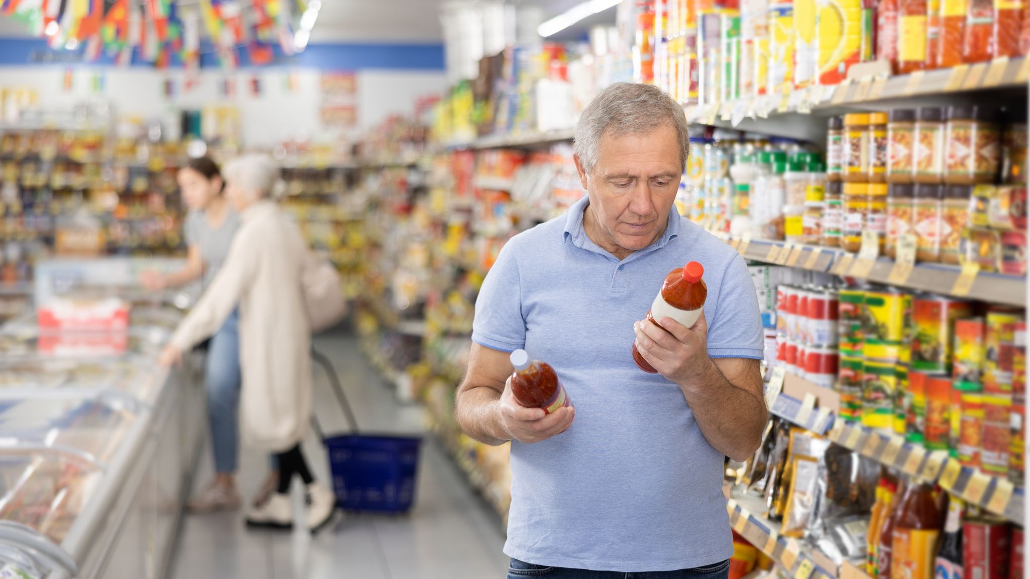 A mature man is reading the labels to compare different brands of hot sauce inside a grocery store.