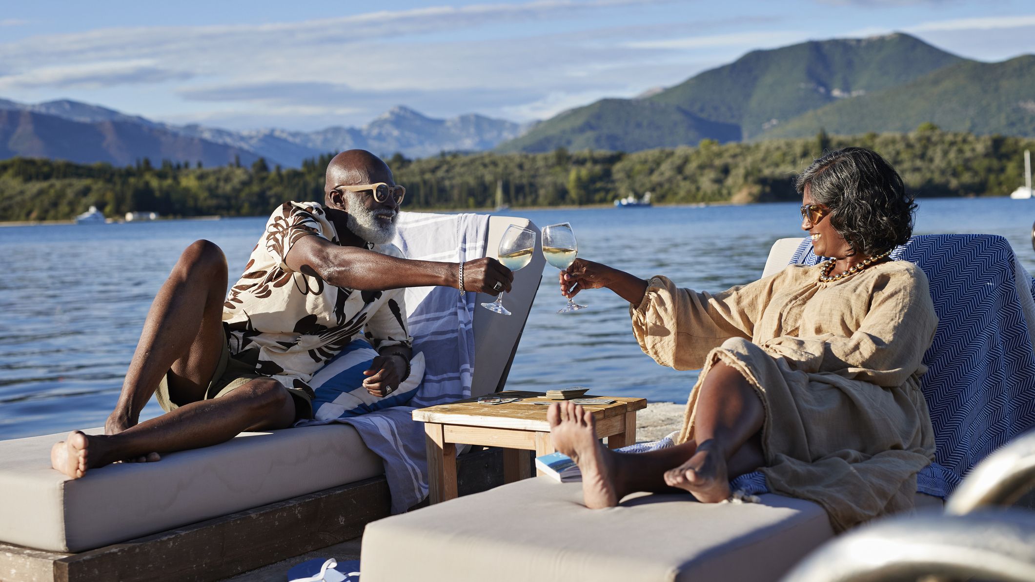 A senior couple clinks glasses while relaxing by a lake.