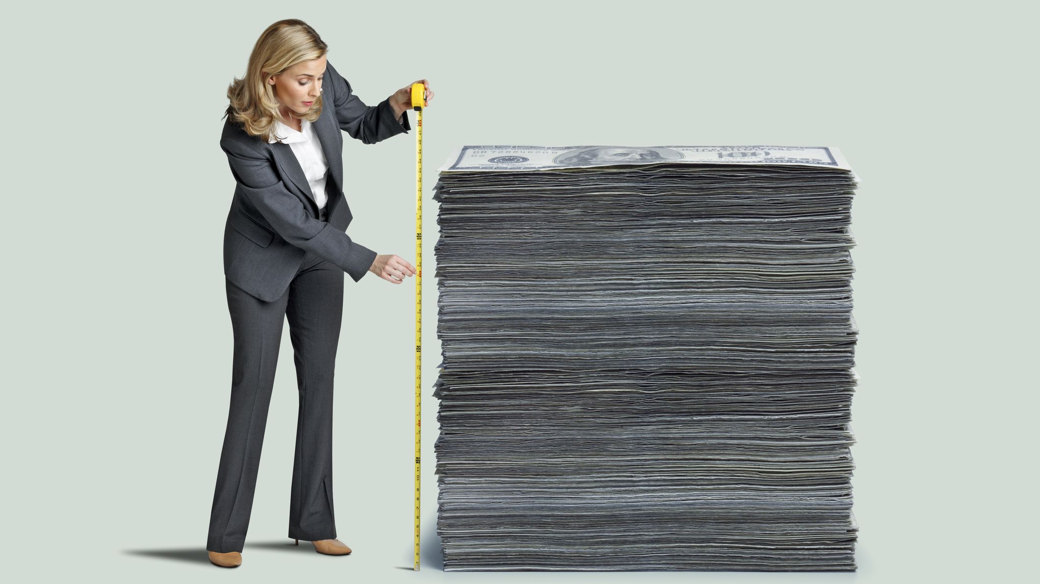 A woman measures a stack of one hundred dollar bills with a yellow tape measure isolated on a green background.
