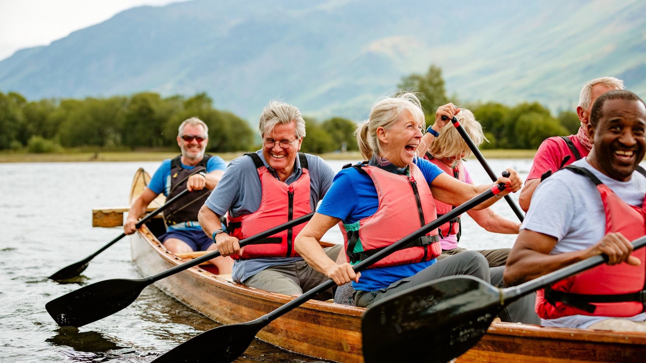A group of older friends laugh as they row a long, wooden canoe.