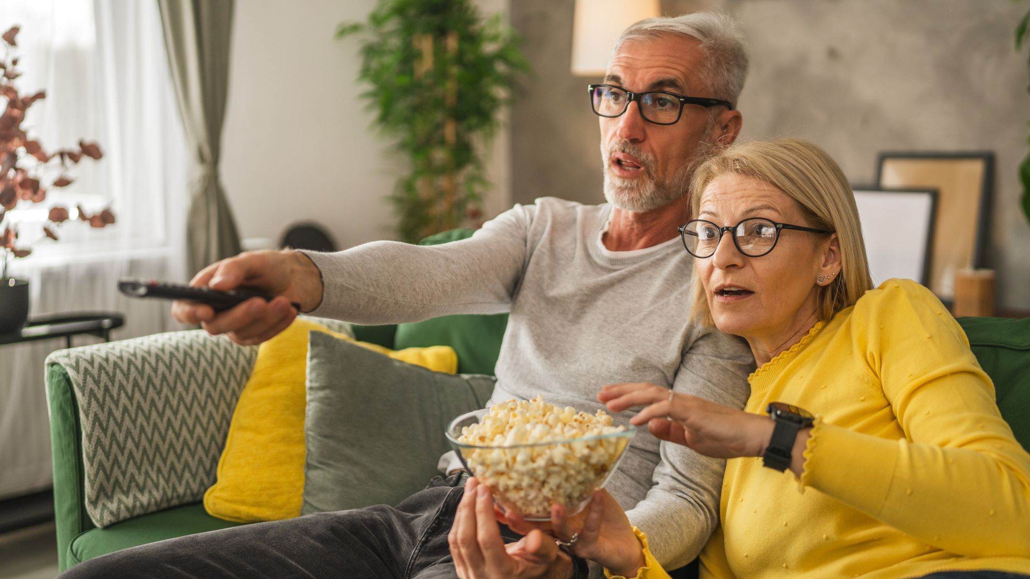 Mature senior couple husband and wife watch scary movie and hold popcorn with remote control at home