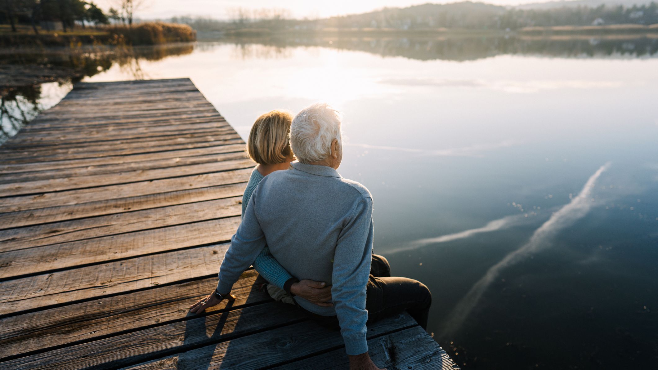 An older couple sits on a dock watching sunrise or sunset over a lake. She has her arm around him, and they face away from the camera.