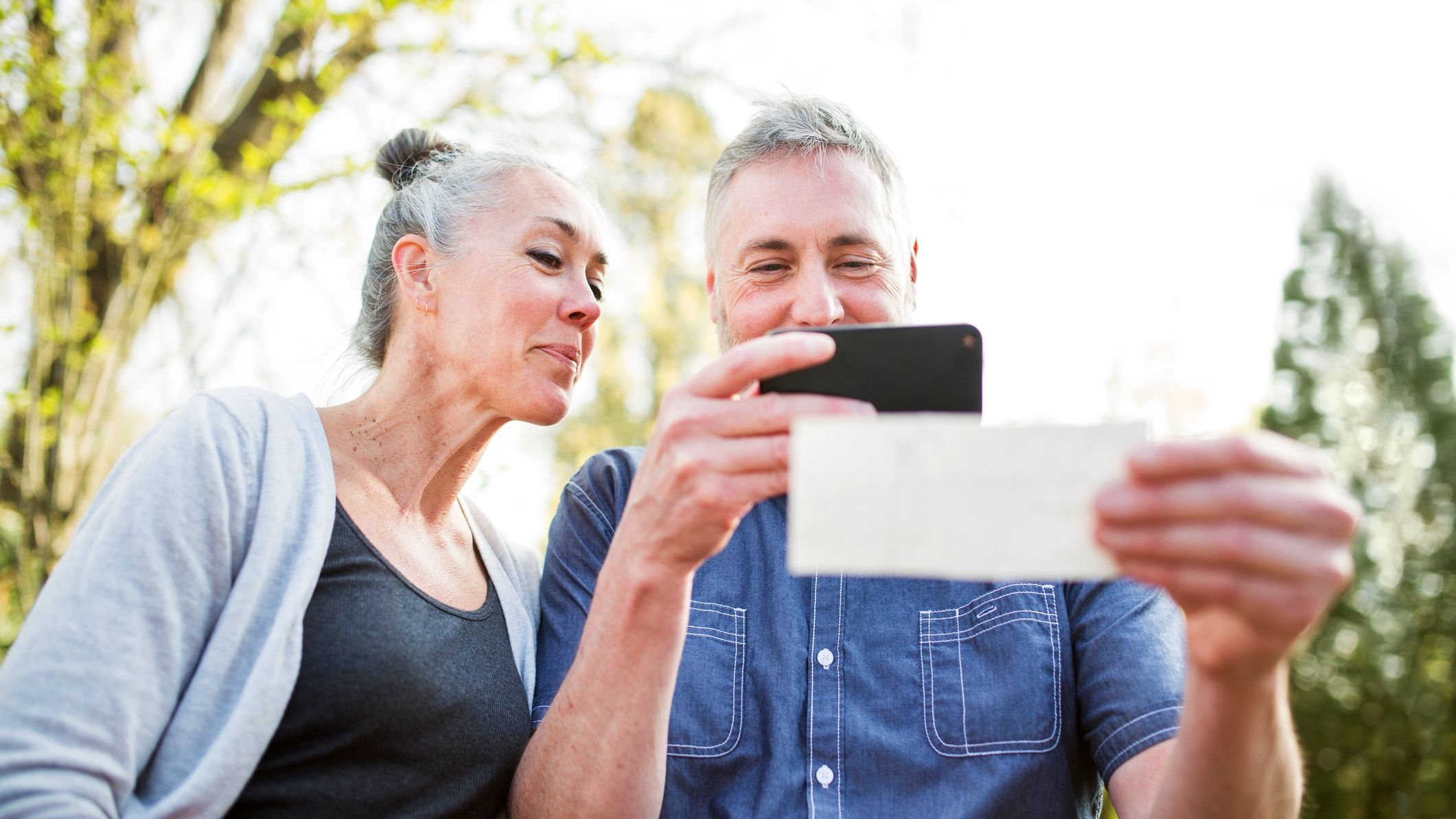 An older couple smiles at a check as they scan it with a cellphone for a direct deposit. The photo may symbolize guaranteed income in retirement.