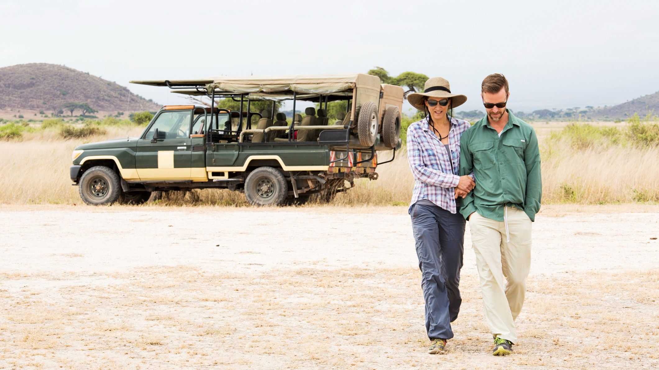 A middle-aged couple in sunglasses walks away from a large safari jeep.