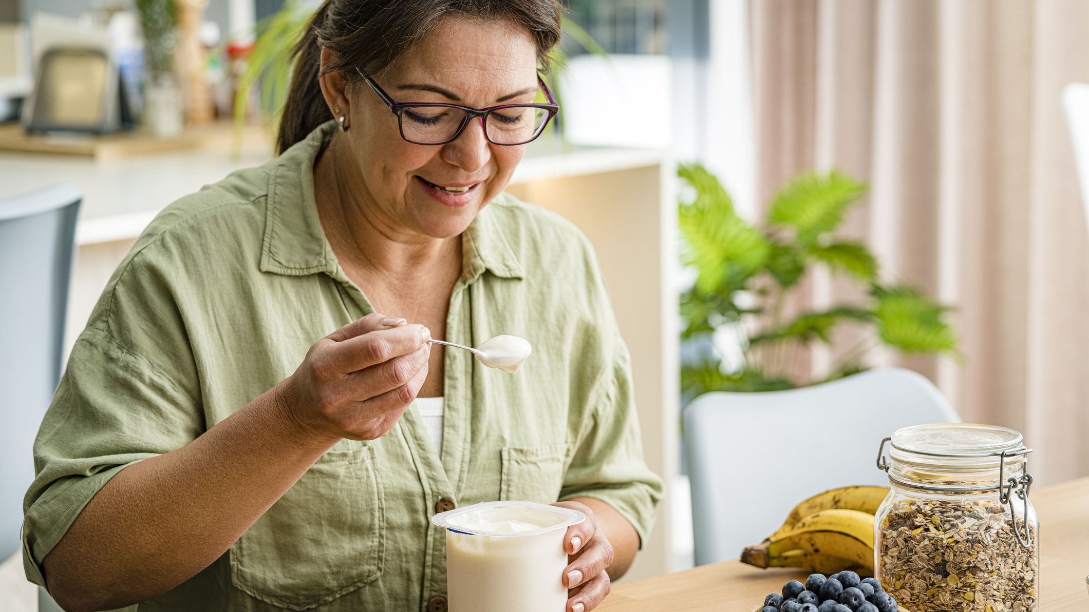 Woman holding spoon of Greek yoghurt, with a bowl of blueberries, a bowl of pumpkins, a container of oats and a bunch of bananas on the counter she&#039;s sitting at