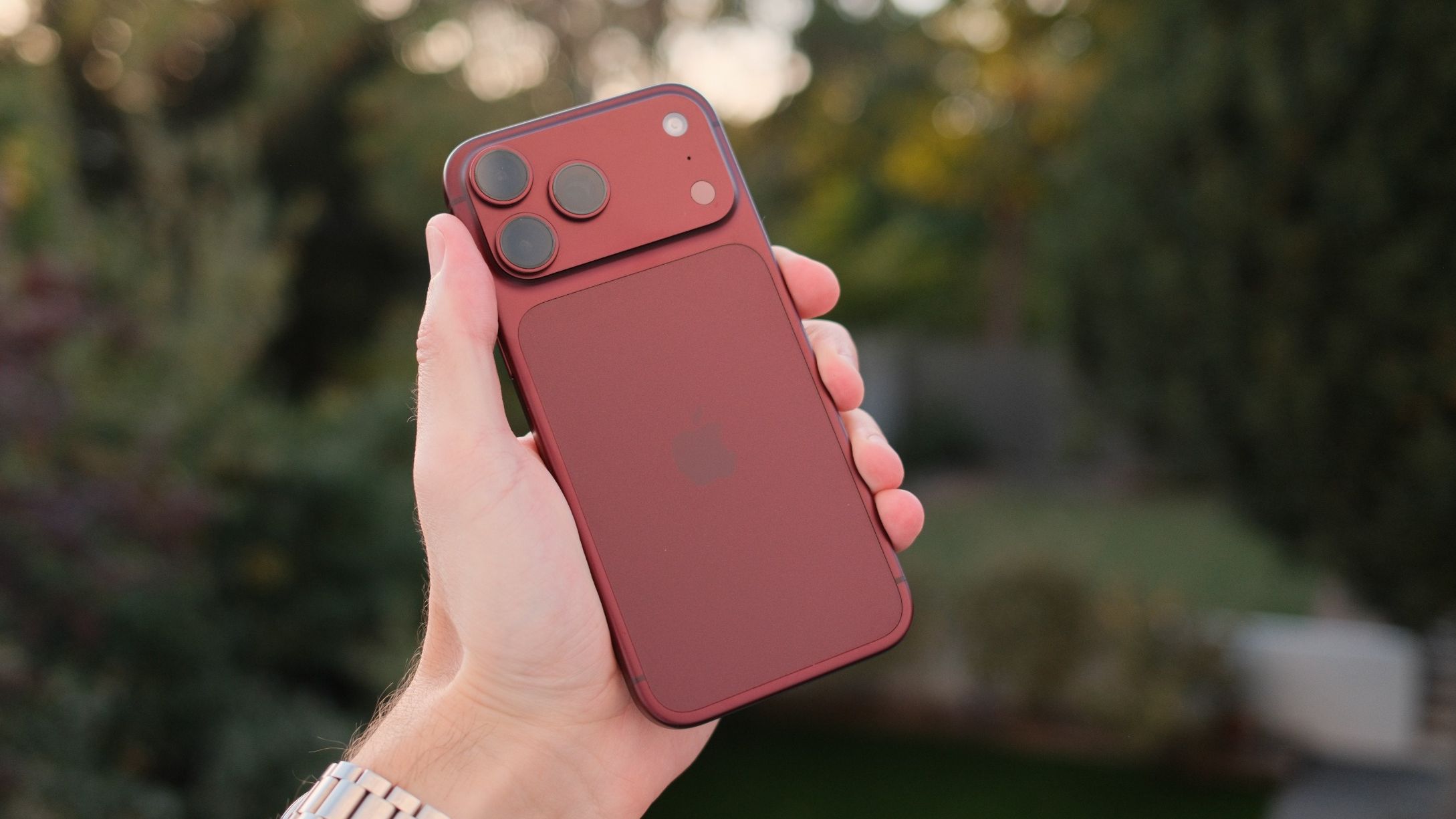 Burgundy iPhone 18 Pro Max held up in a garden, showing the rear design and triple-camera system.