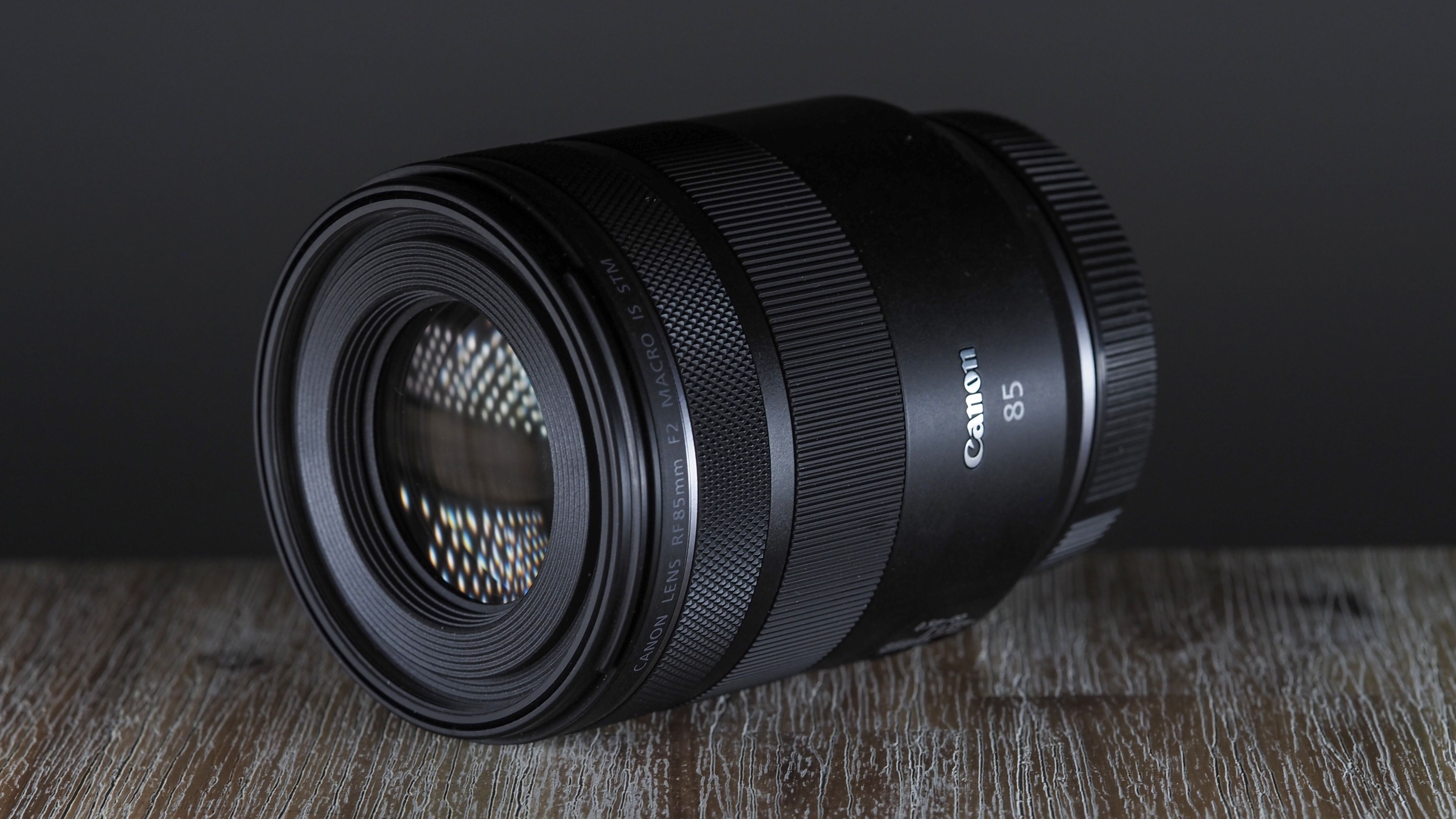 Canon RF 85mm f/2 Macro lens on a table, against a dark backdrop