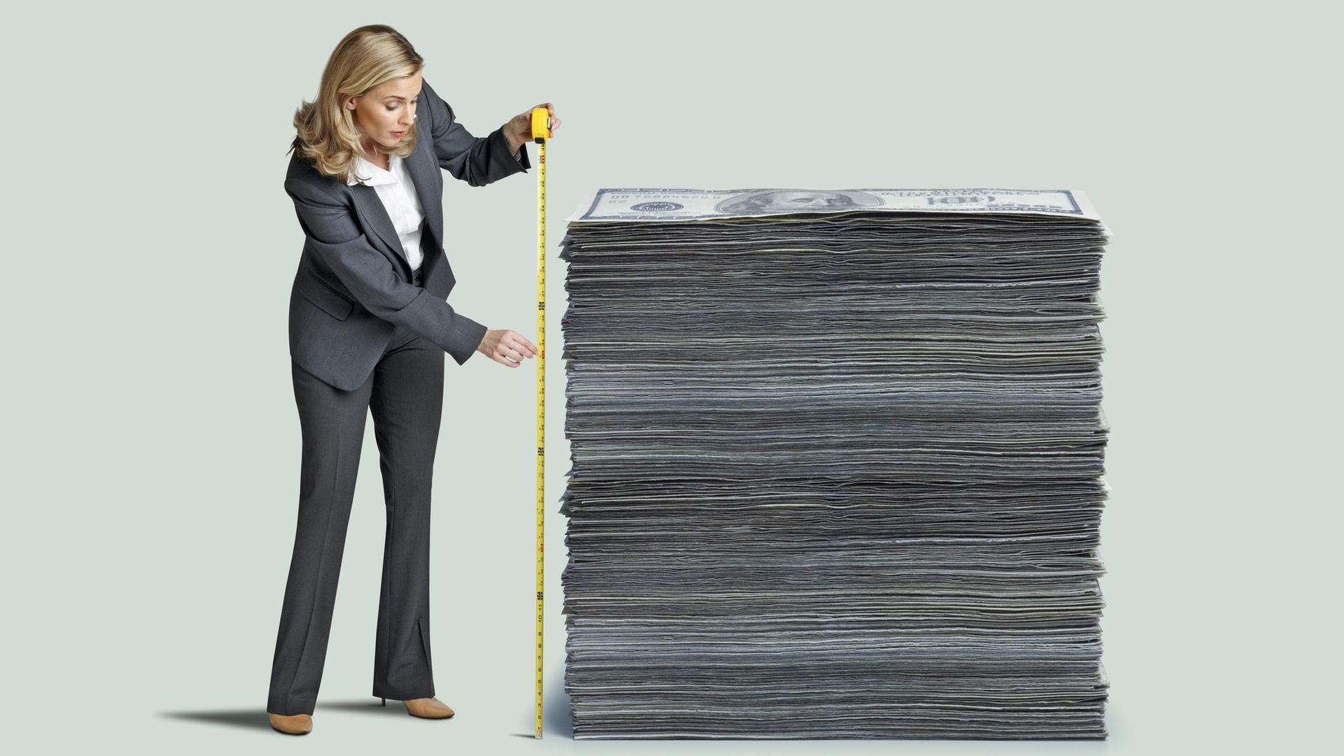 A woman measures a stack of one hundred dollar bills with a yellow tape measure isolated on a green background.