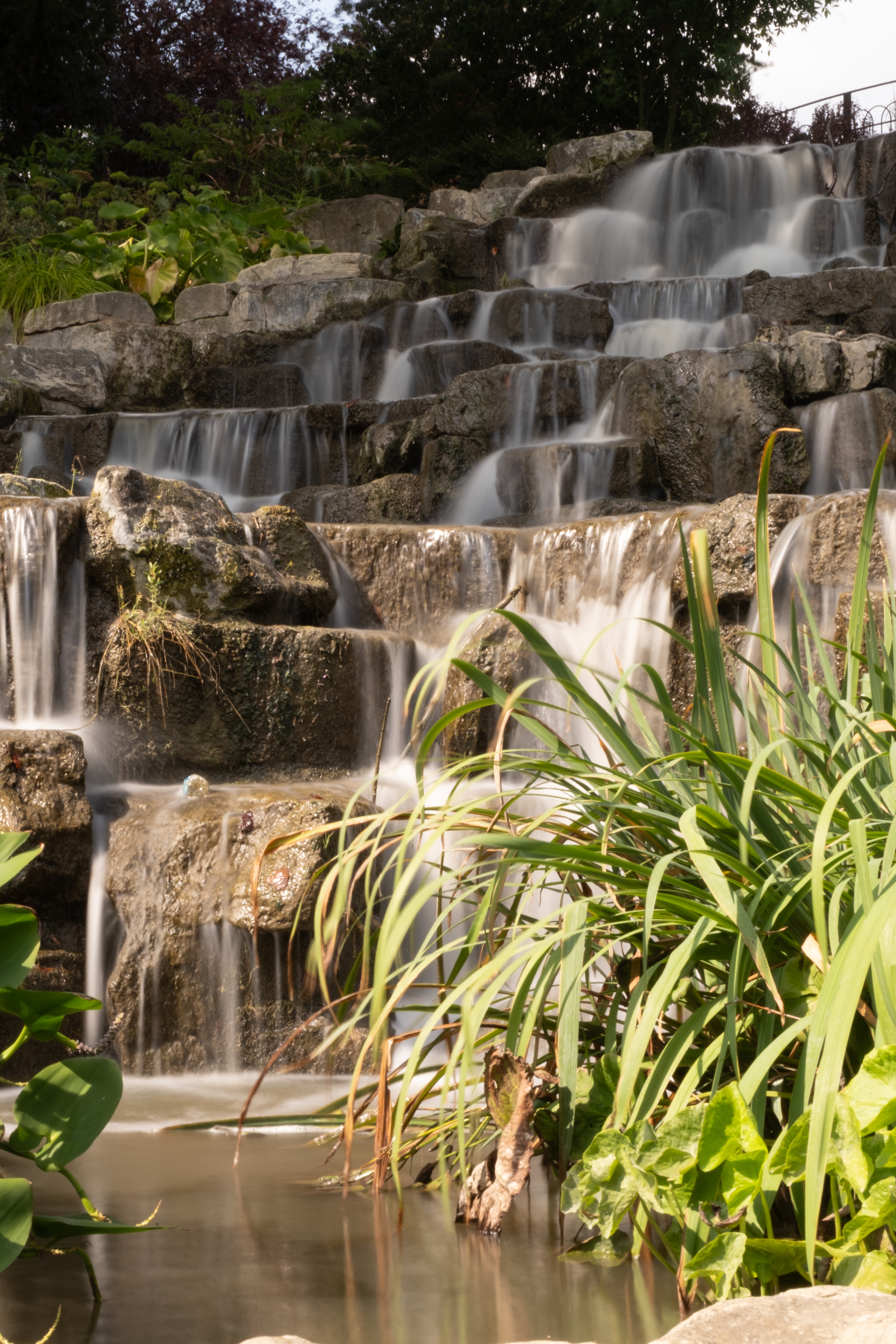 Long-exposure photograph of a tiered rock waterfall surrounded by green plants, with water rendered as smooth, flowing streaks.