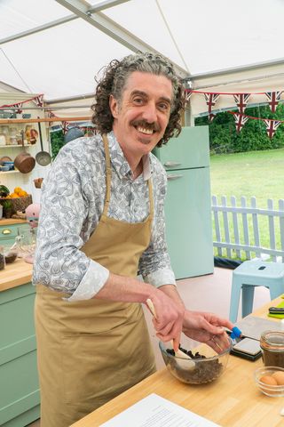 Giuseppe in a grey shirt and beige apron stands in the tent stirring food in a bowl on The Great British Bake Off.