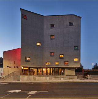 Alternative exterior view of the grey concrete Pálás Cinema building with the lights on during the evening. The building features multiple windows and the wording 'PALAS'. Directly outside the building is a ramp leading up to it and a road